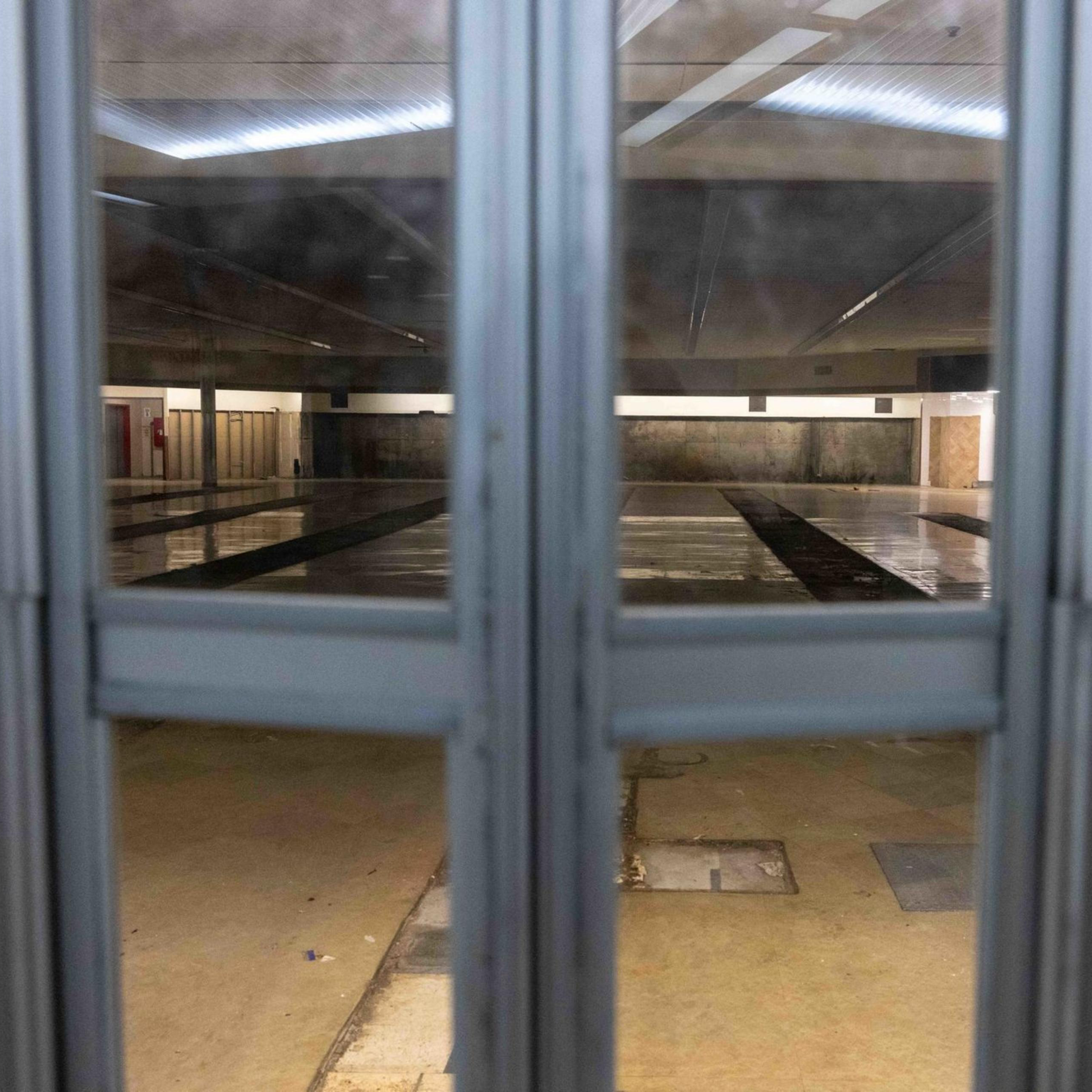 The image shows an empty indoor swimming pool, viewed through a set of glass doors, with lanes visible on the pool floor and overhead lighting.