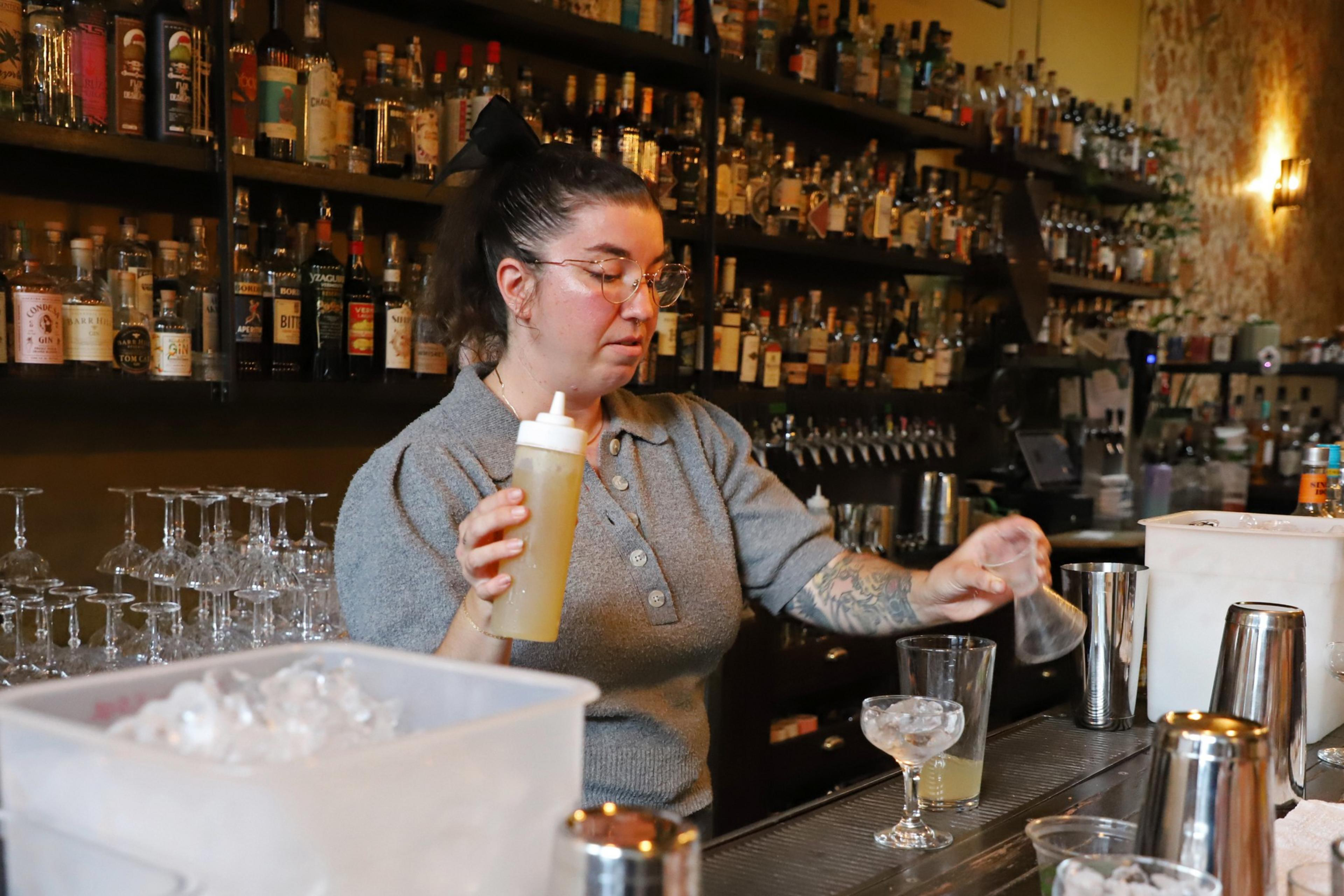 A bartender with glasses and a tattoo pours a yellow liquid from a squeeze bottle into a mixing glass at a well-stocked bar.
