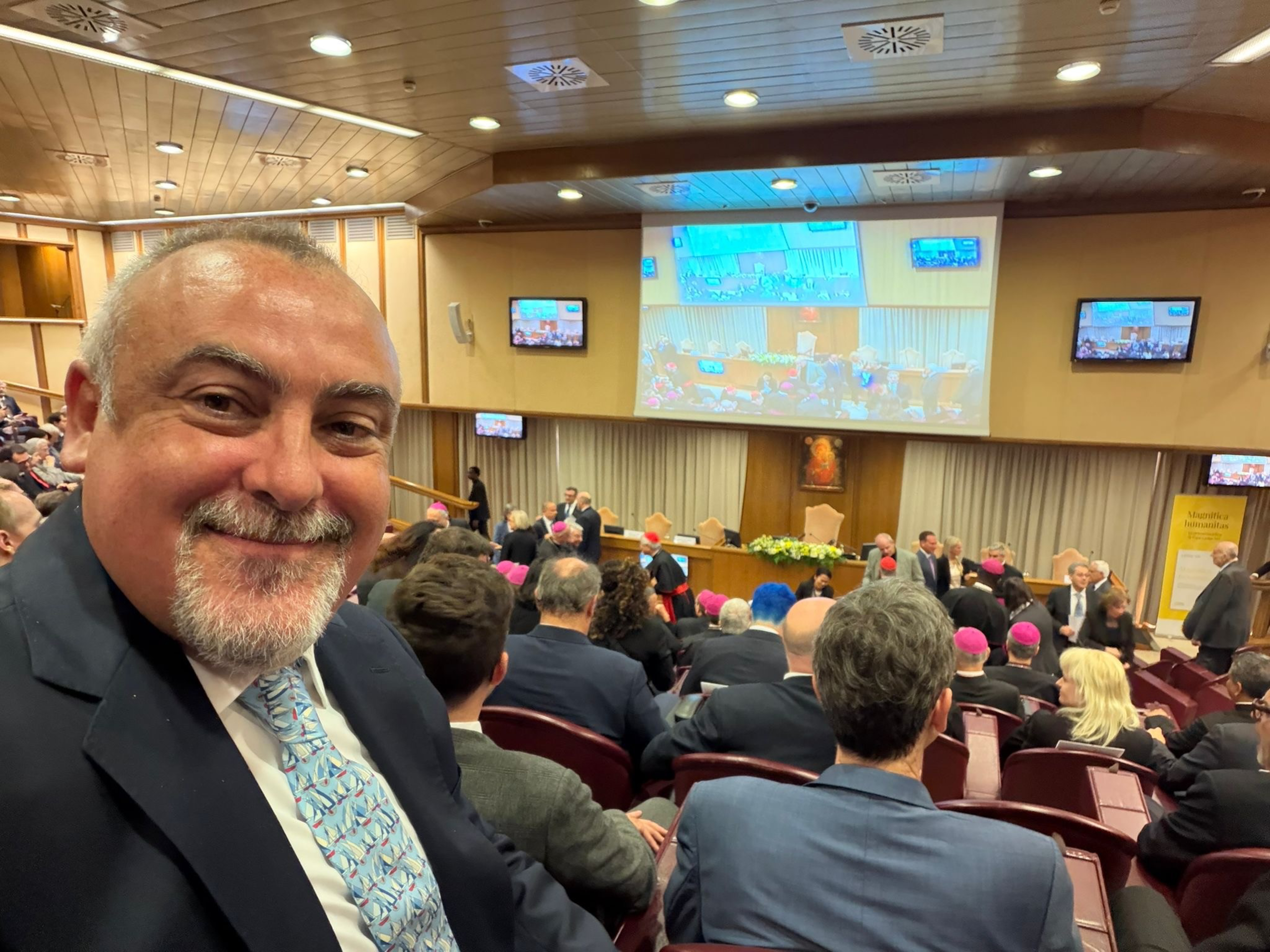 A man in a suit smiles at the camera inside a conference room filled with seated people, some wearing pink skullcaps, and a large screen at the front.