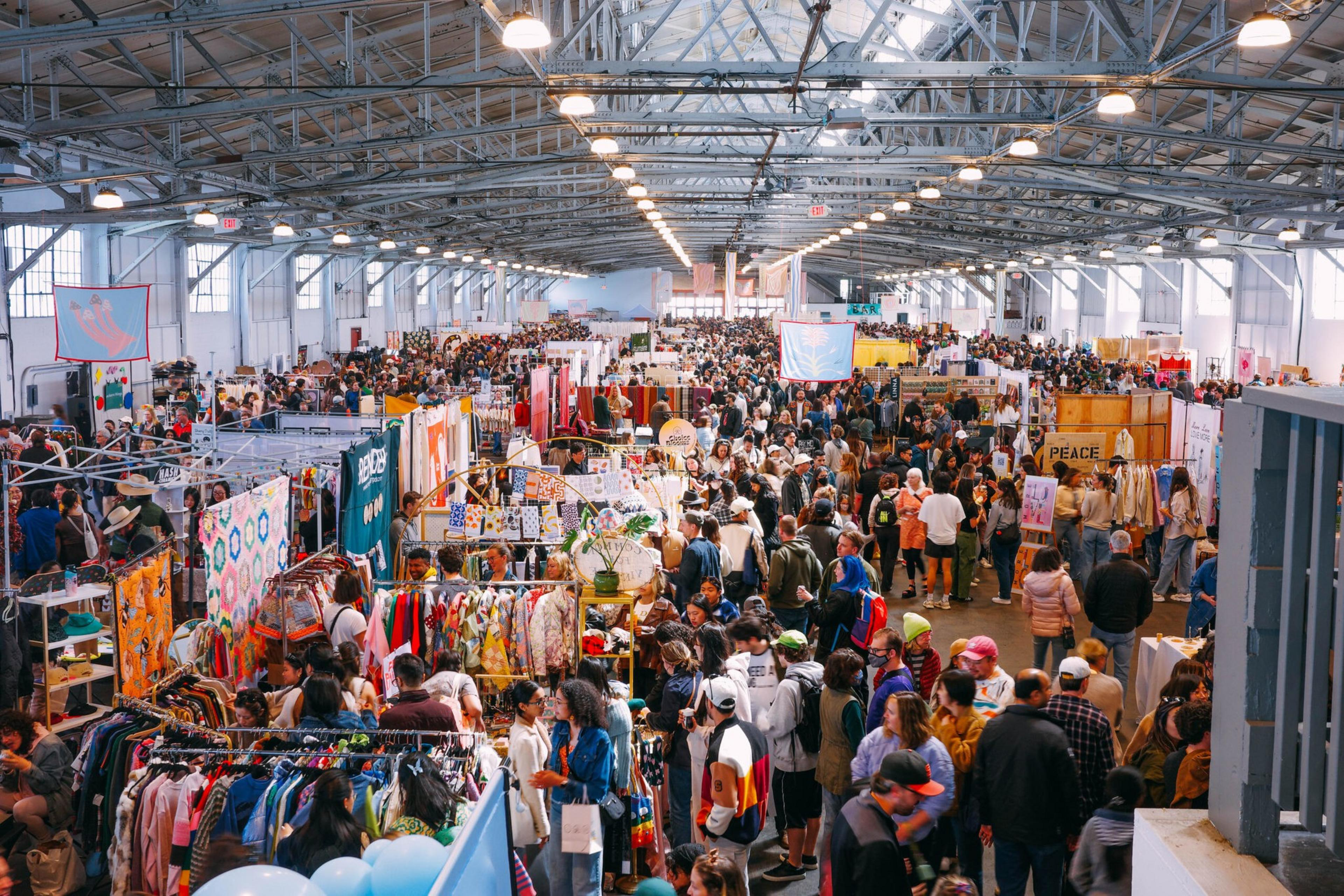 The image shows a crowded indoor market with people browsing colorful booths under a high, industrial-style ceiling, creating a lively and bustling atmosphere.