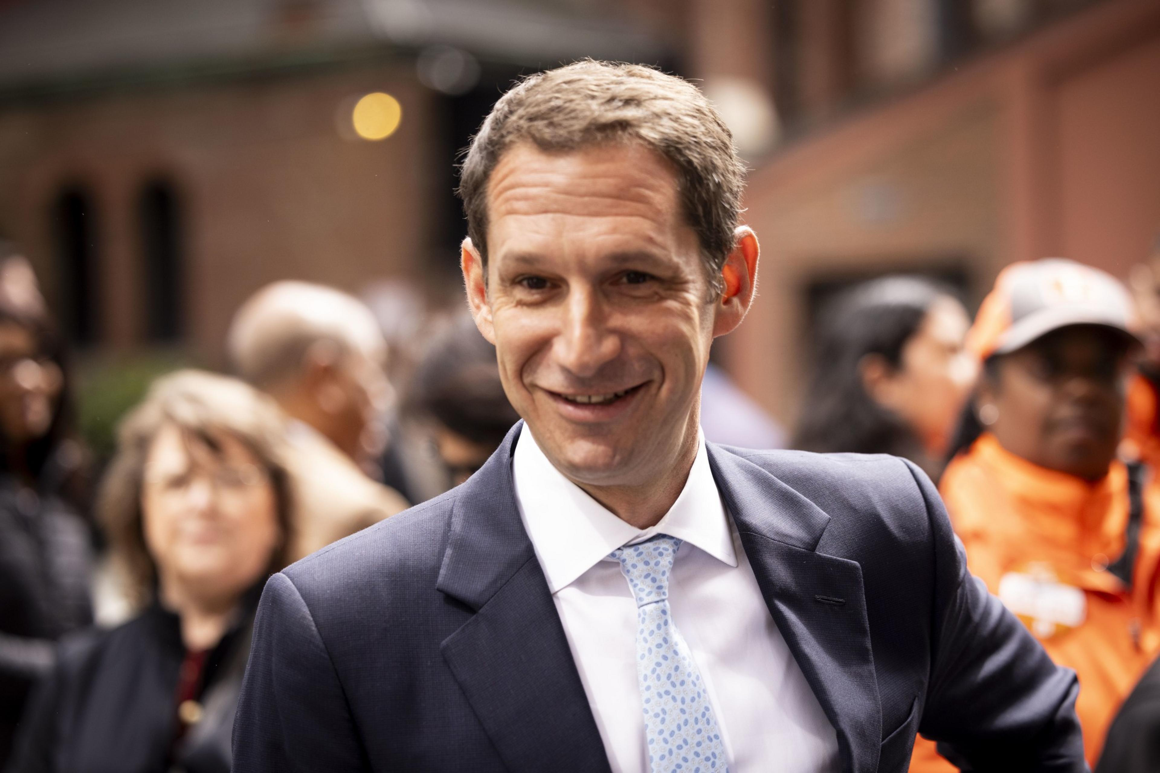 A man in a suit and tie smiles outdoors, surrounded by a crowd with blurred faces in the background.