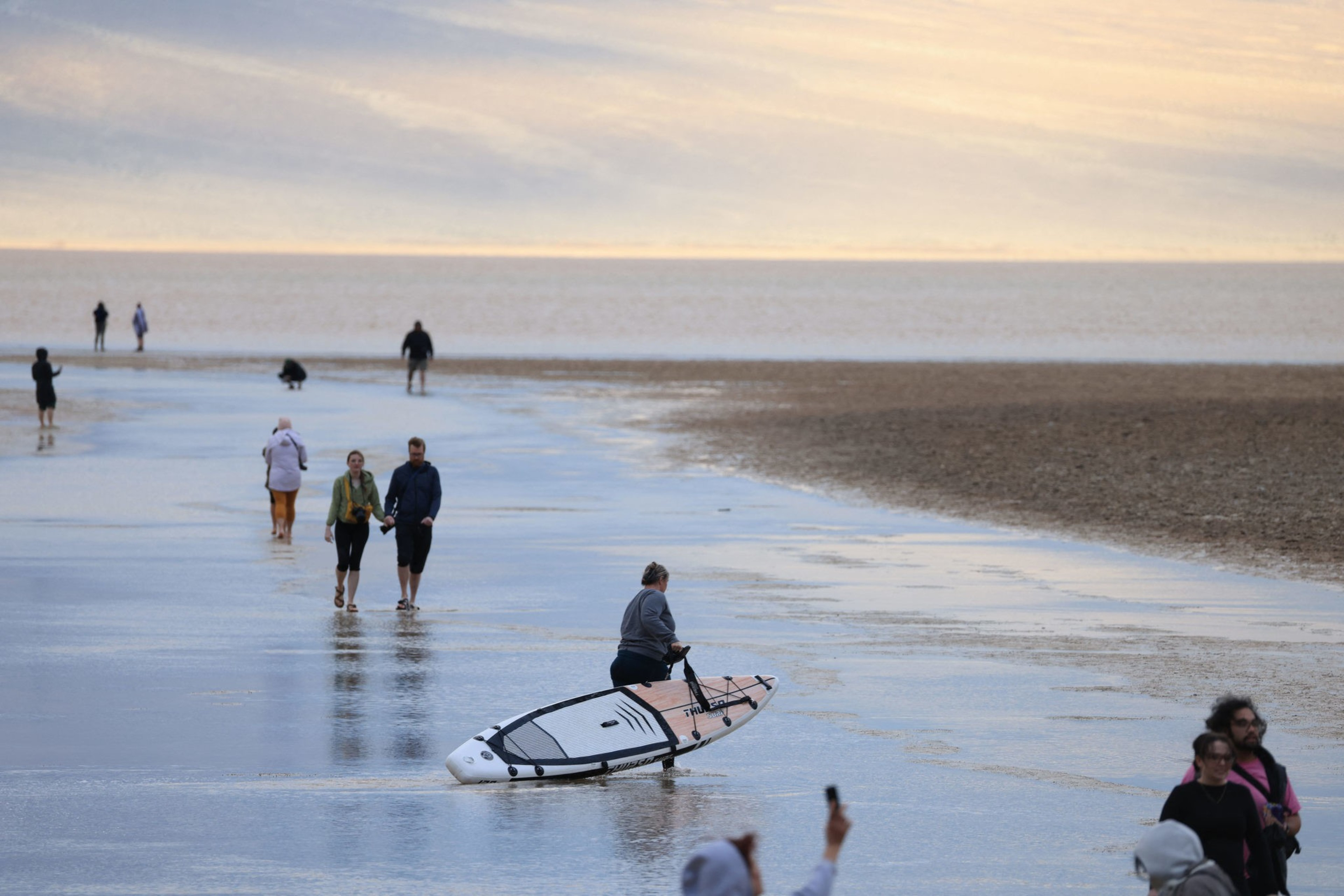 shot from afar, people wade through a shallow lake by day, with one person carrying a small boat