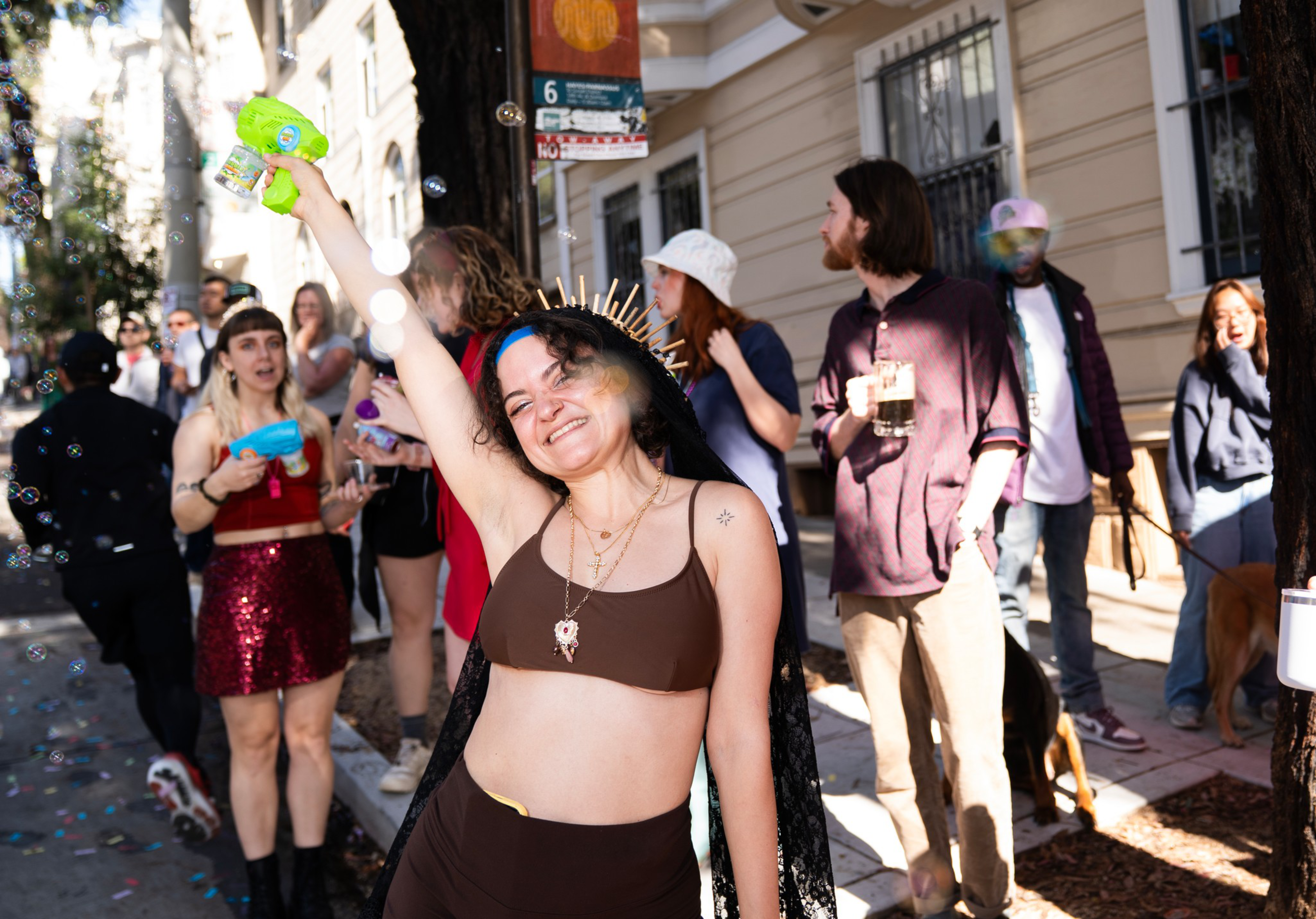 A smiling woman in a brown outfit and golden headpiece sprays bubbles with a green toy gun among a lively crowd on a sunny street.