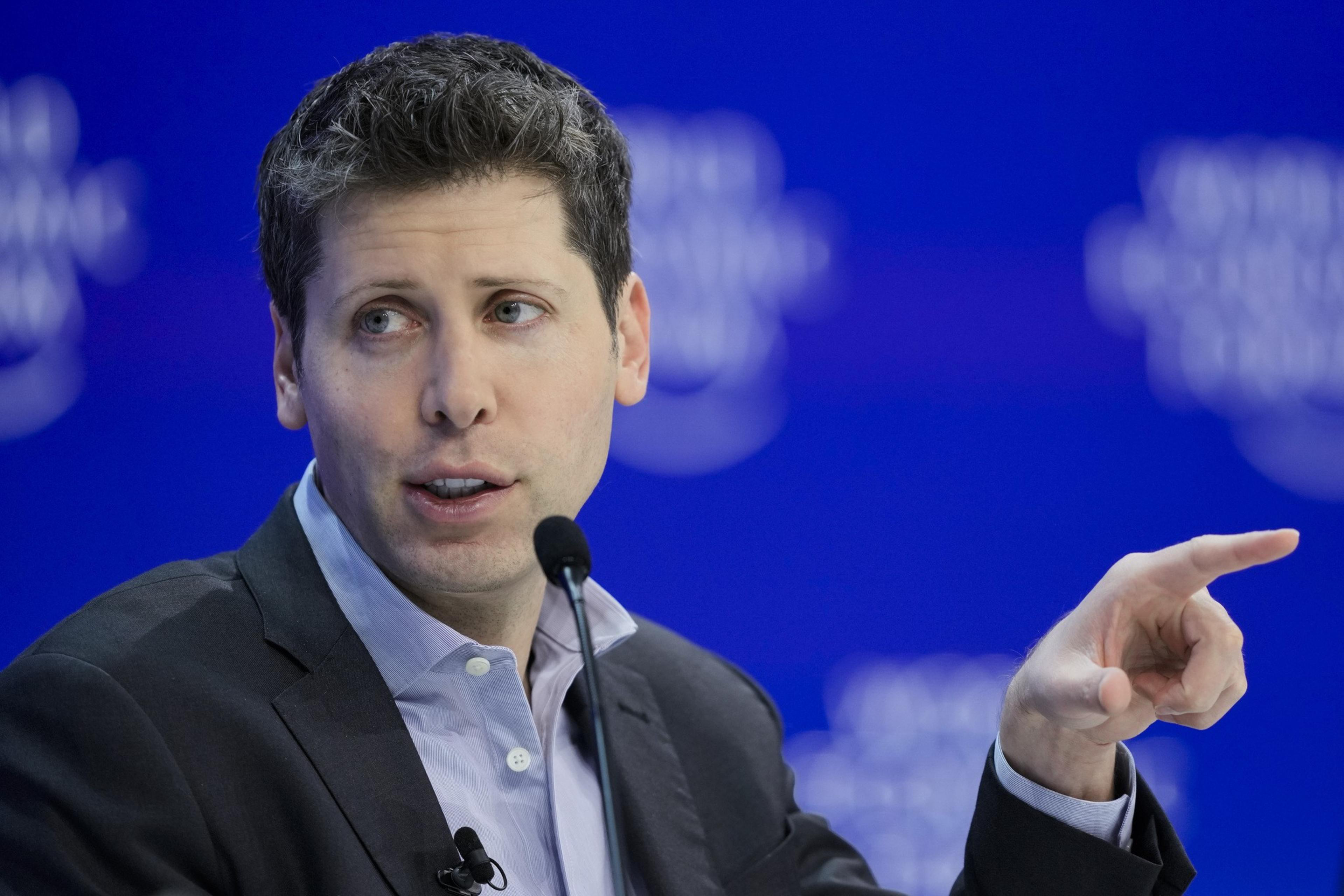A man gestures while speaking, microphone nearby, against a blue backdrop with blurred circular lights.