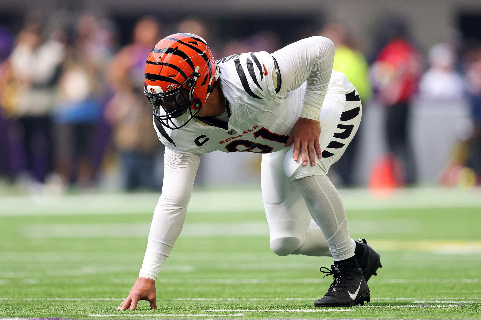 A football player in a white uniform with black tiger stripes and an orange helmet crouches on the field, ready to start a play.