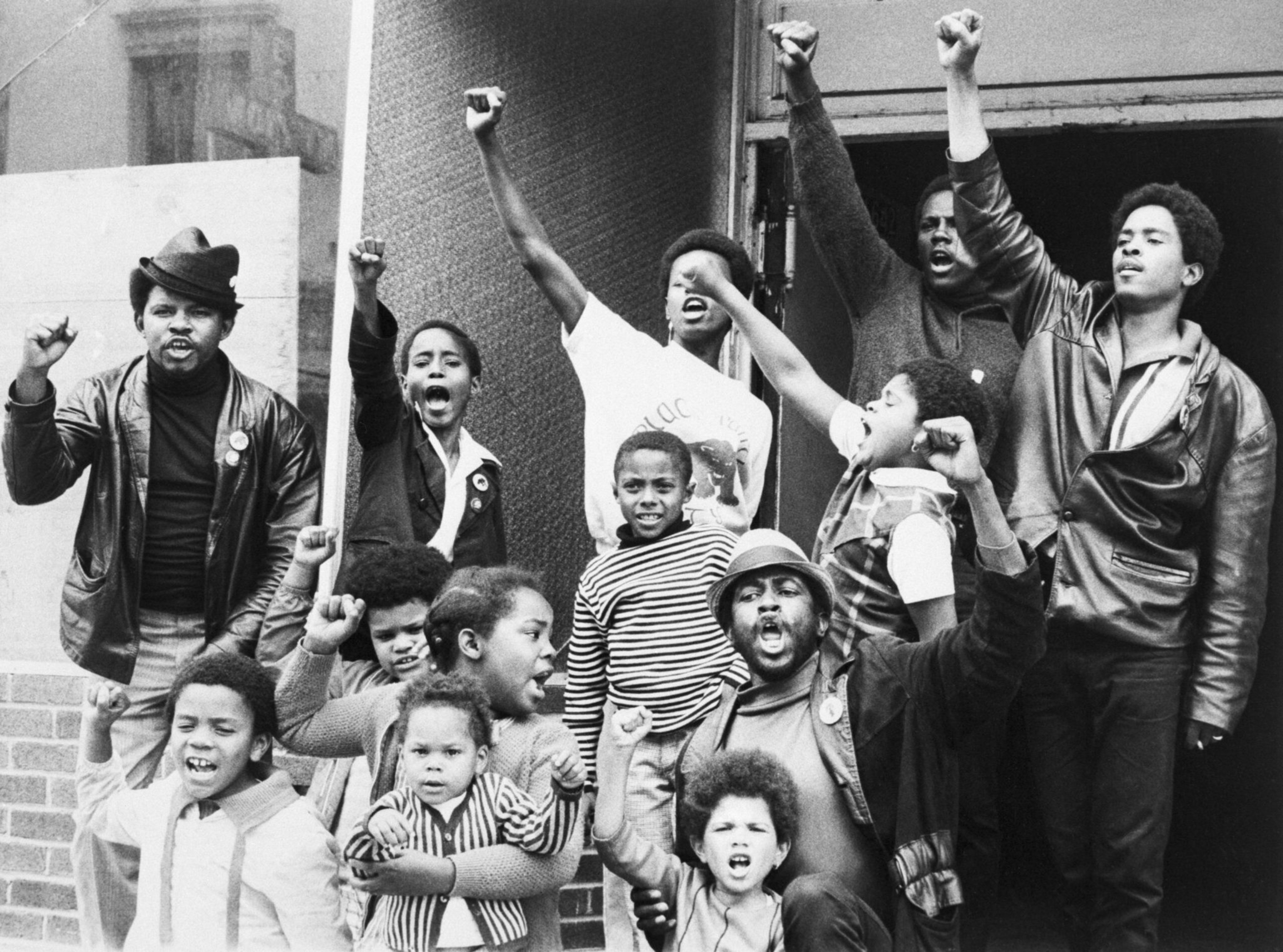 A diverse group of Black people, including children and adults, raise their fists in solidarity, expressing unity and strength outside a building.