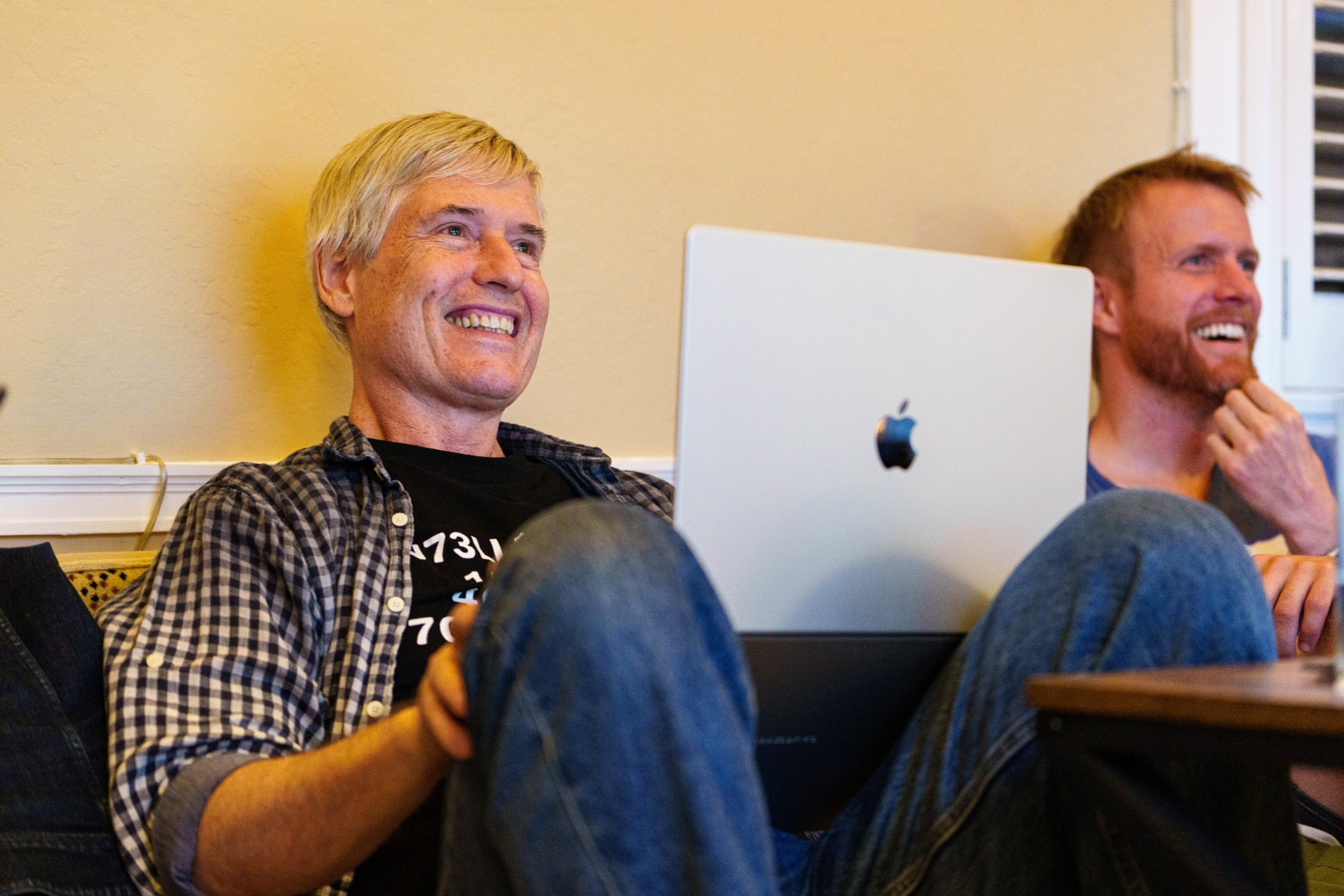 Two men sit close together on a couch, smiling and laughing, with a laptop open on the lap of the man in the foreground.