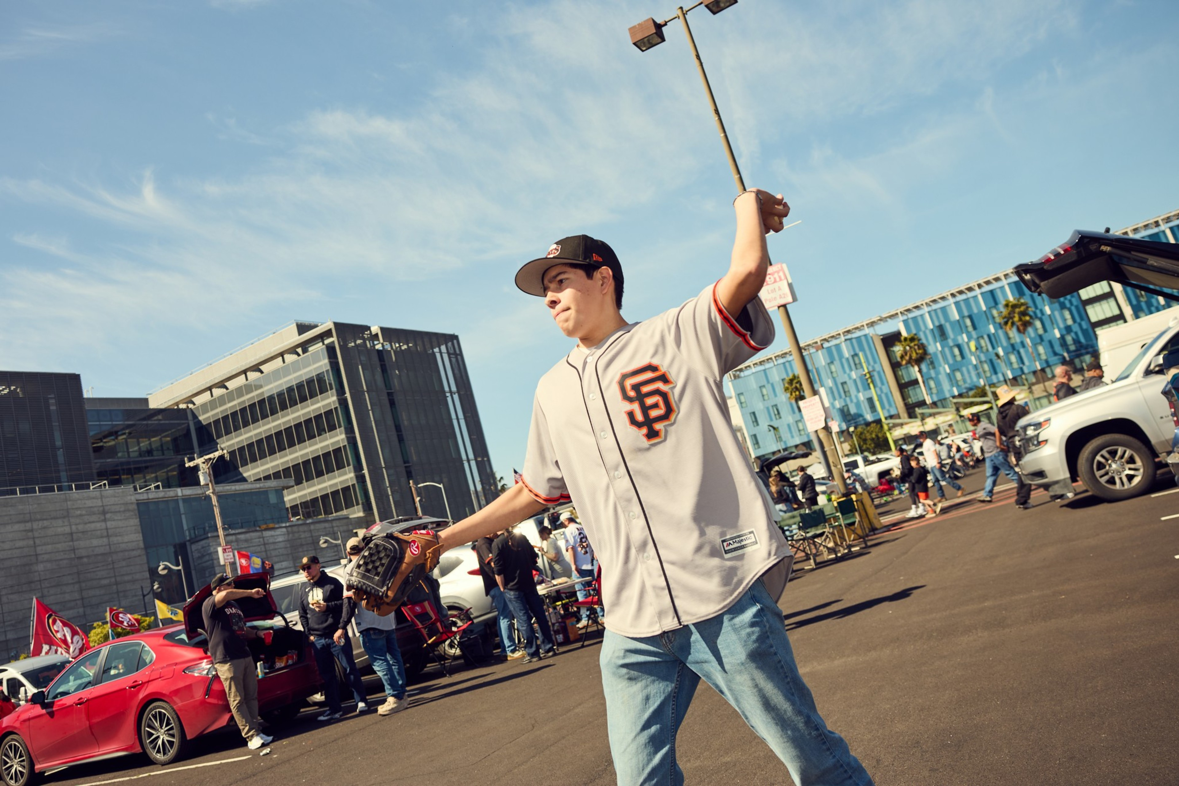 A person in a baseball jersey and cap is playfully throwing a baseball in a parking lot, with cars and a few people in the background.