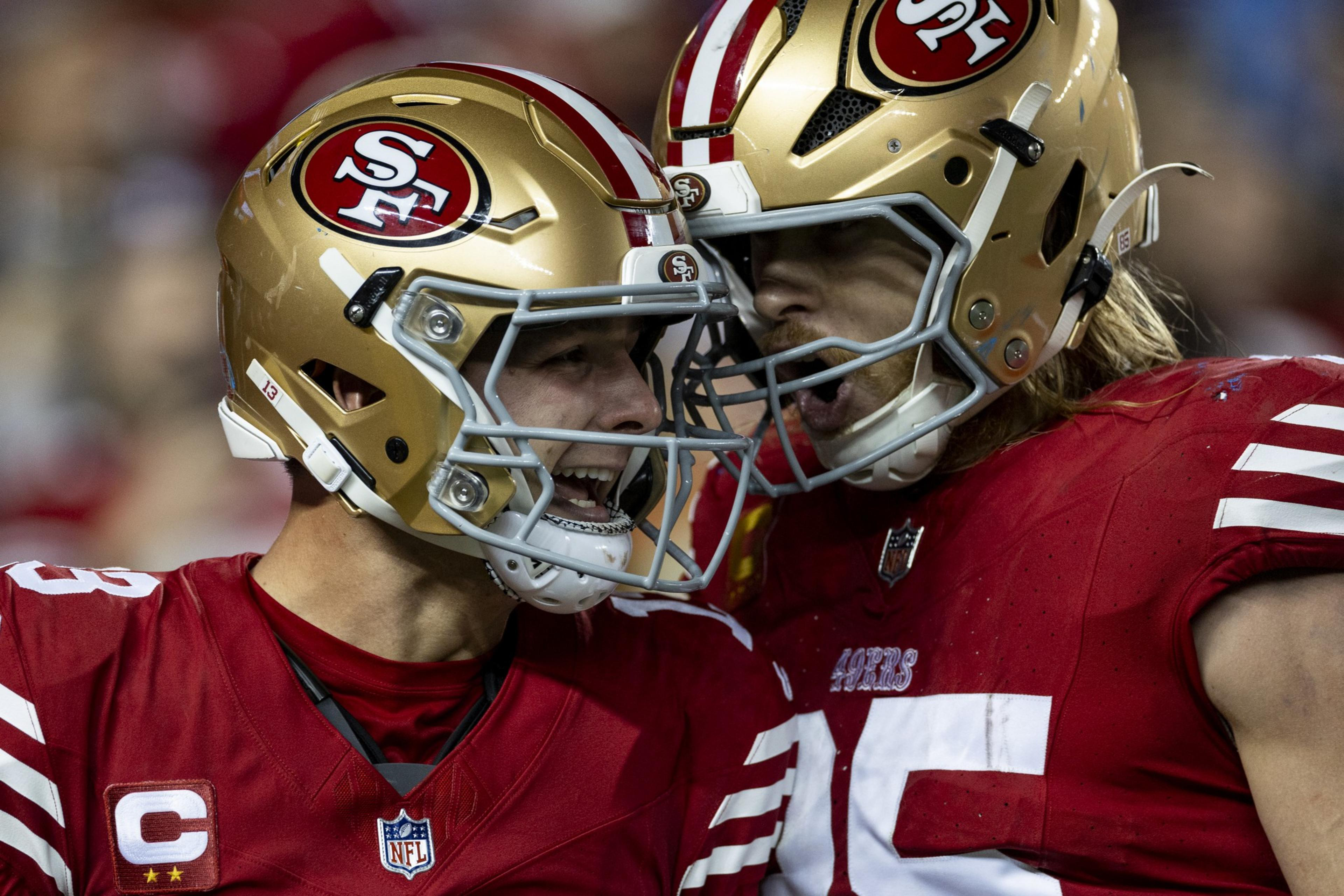 Two football players in red jerseys and gold helmets are celebrating energetically on the field. They are facing each other with intense expressions.