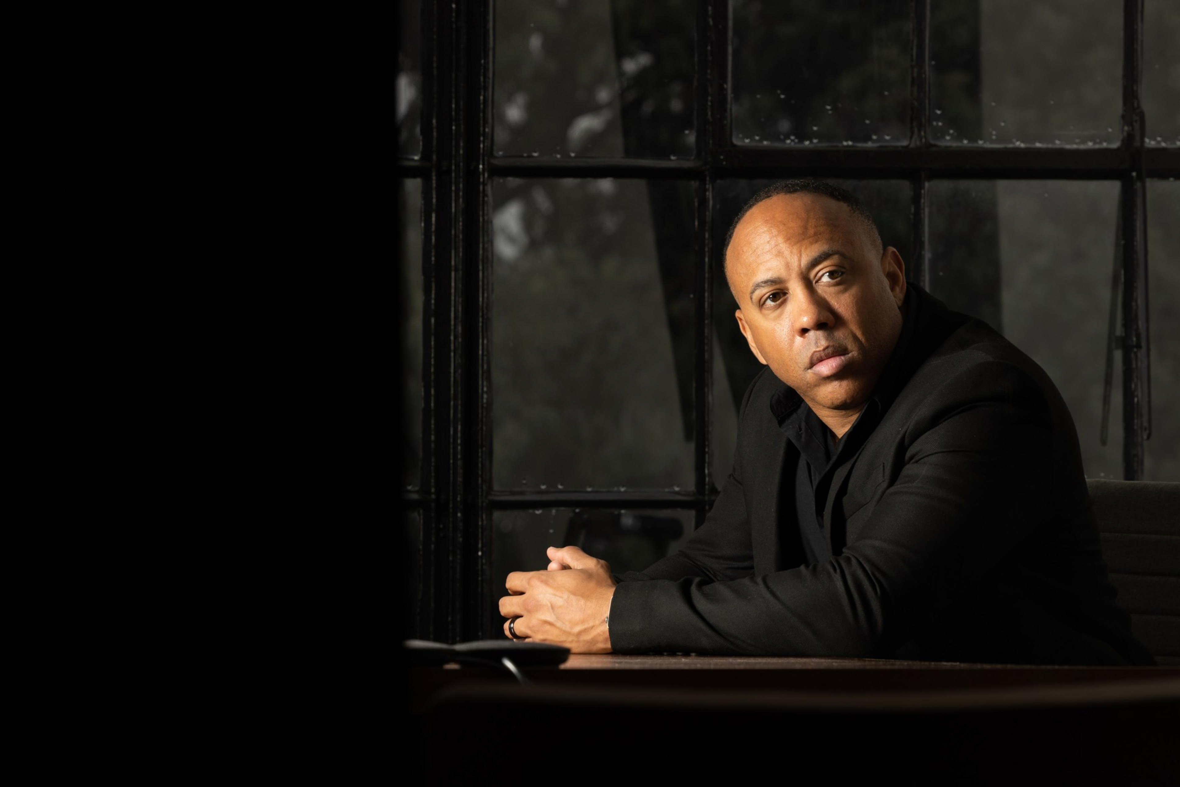 A man in a black suit sits at a table with hands clasped, looking thoughtfully to the side by a large window with a dark, blurred background.