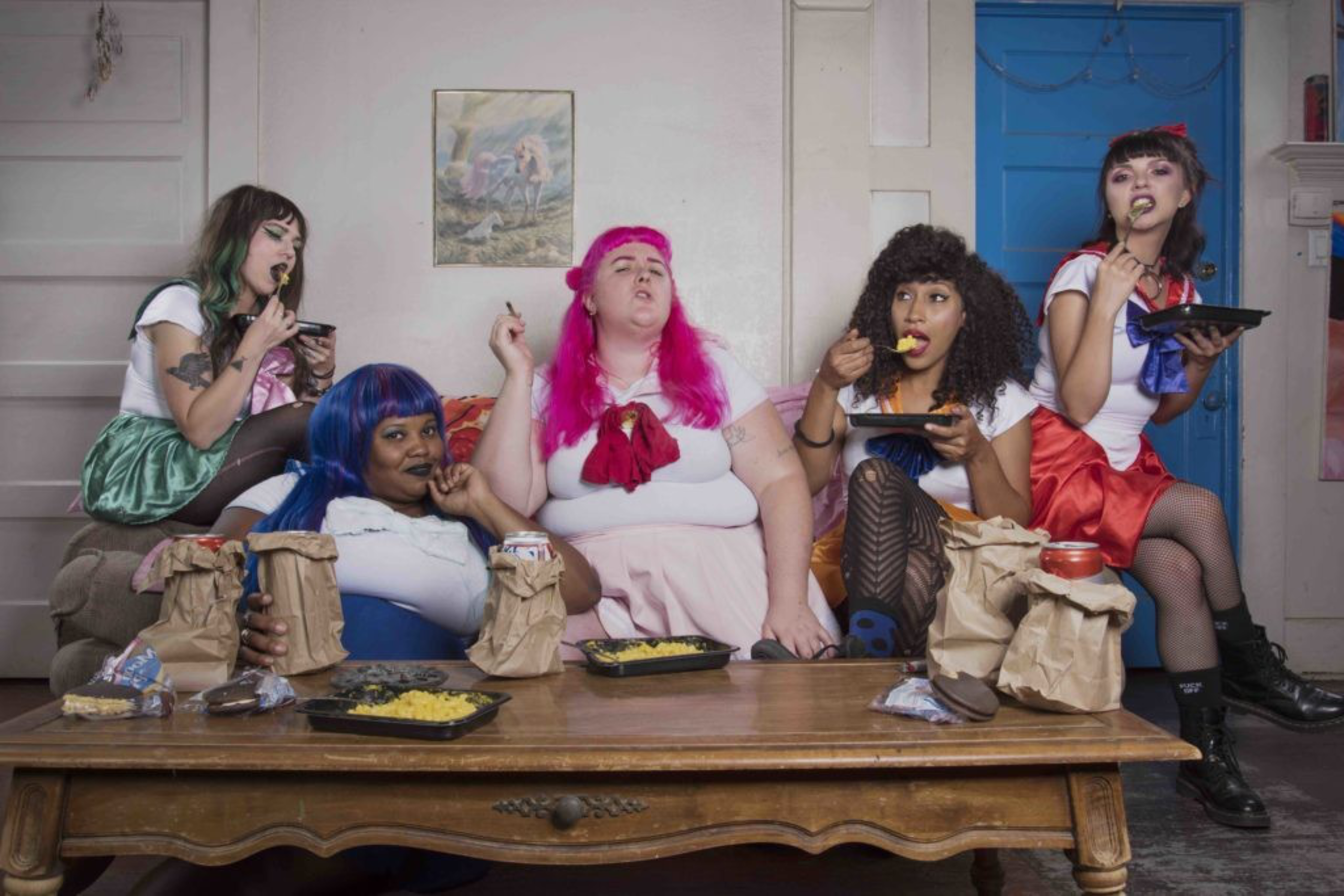 Five women dressed in colorful sailor-style outfits sit around a wooden table, eating from takeout containers and brown paper bags.