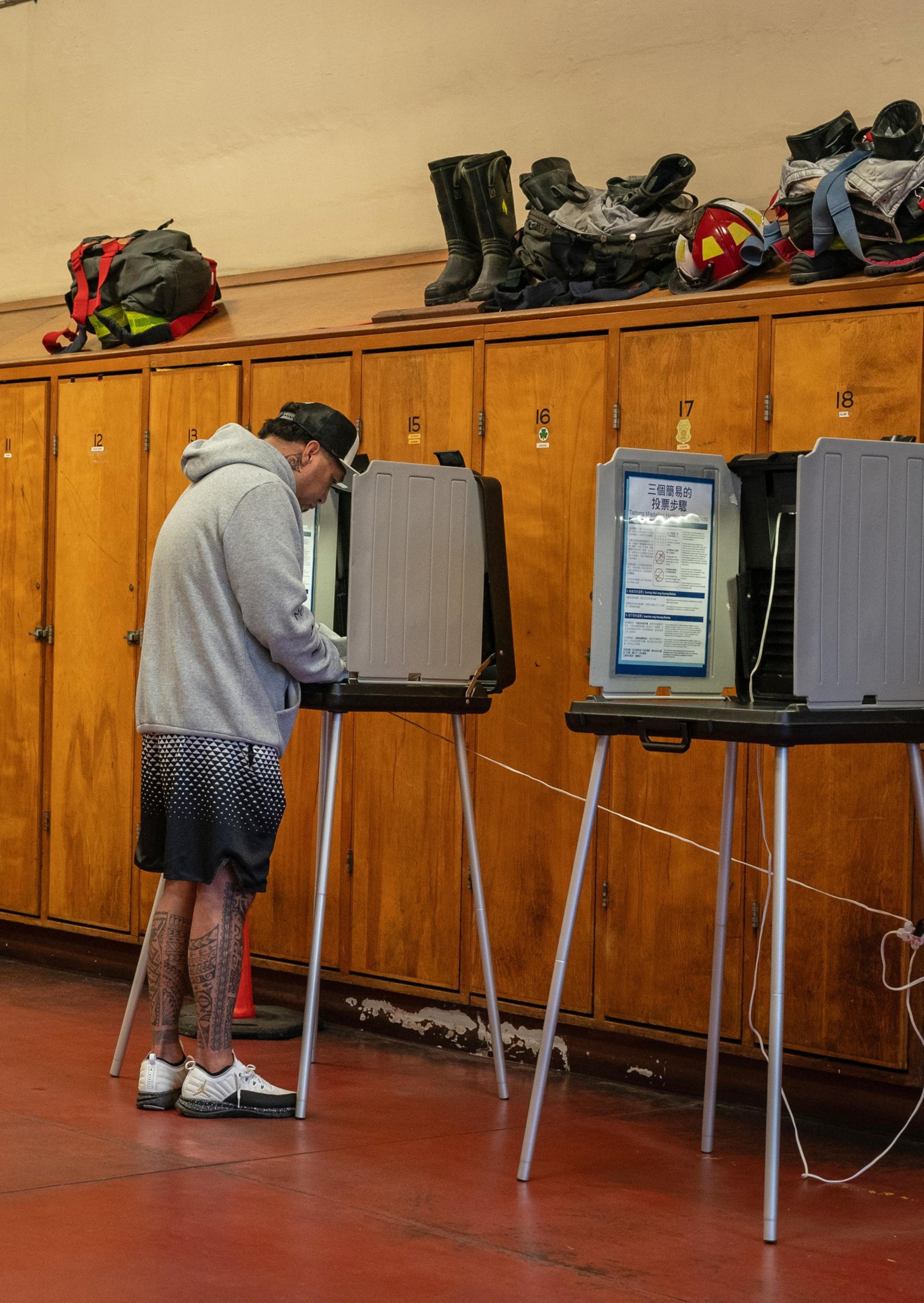 A person in a hoodie and shorts is voting at a booth in front of wooden lockers with gear and helmets on top.
