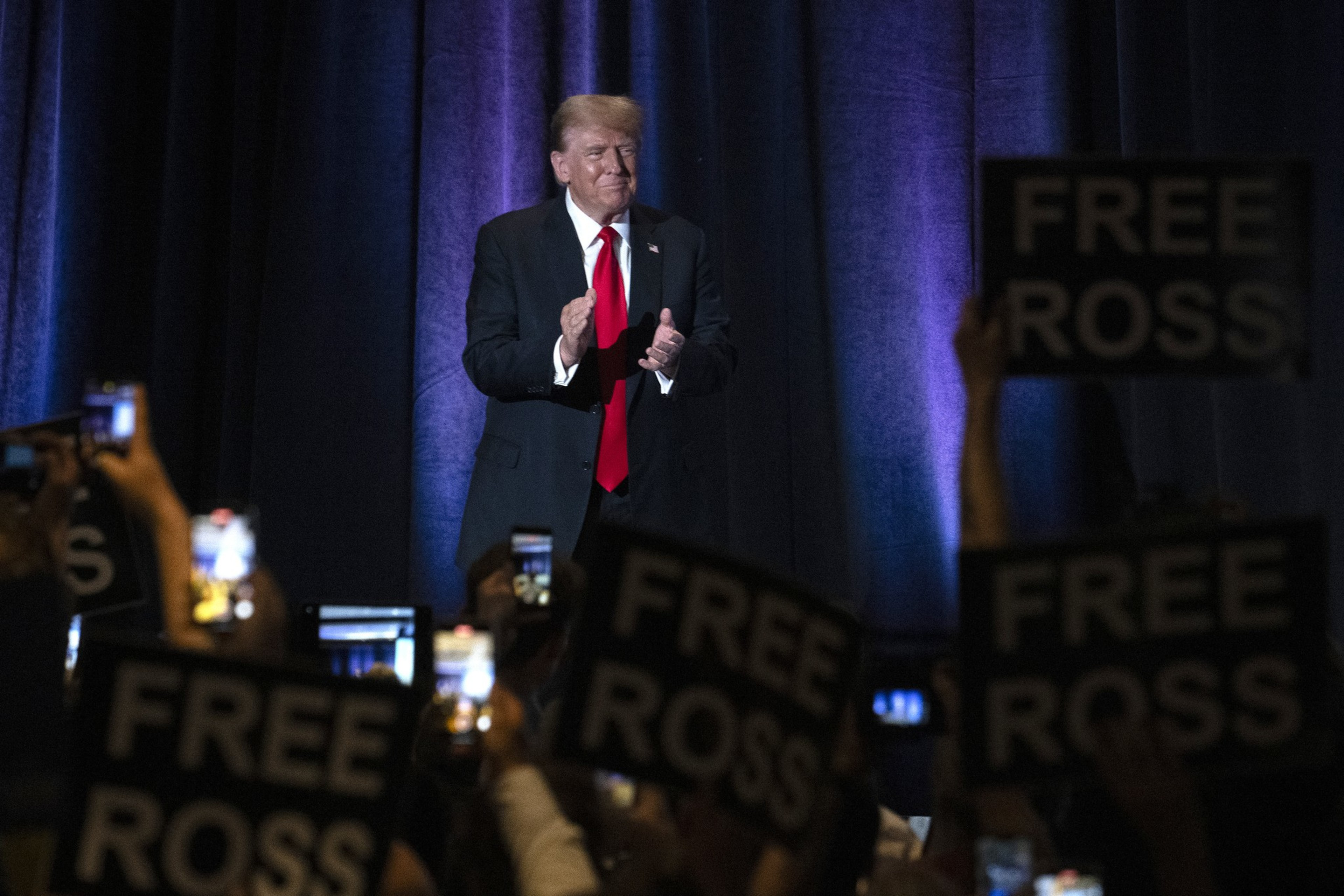 A man in a suit with a red tie claps on stage against a backdrop. The crowd holds signs reading &quot;Free Ross&quot; and takes photos with phones.