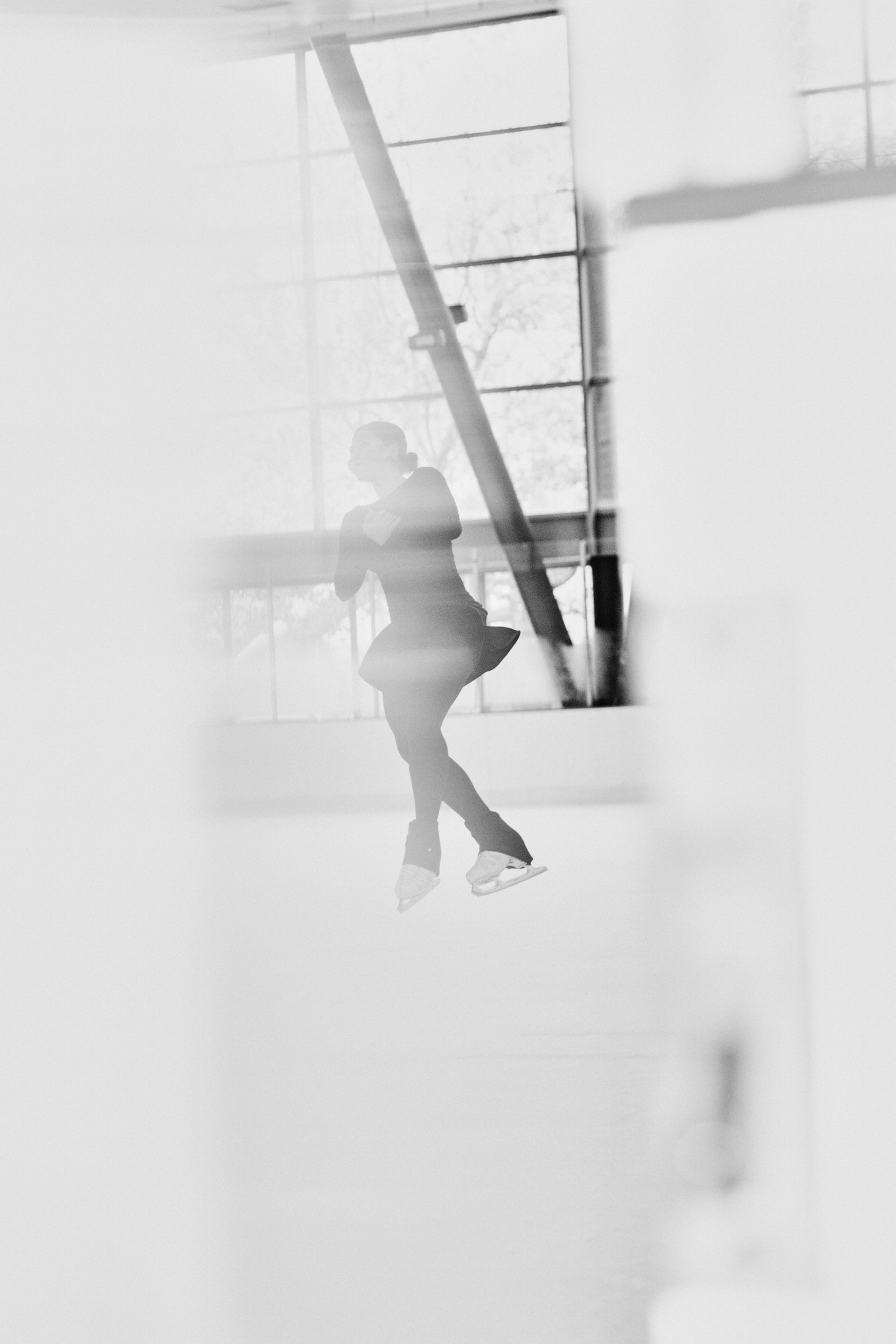 A figure skater mid-jump on an ice rink, captured in black and white with a soft, blurred effect and large windows in the background.