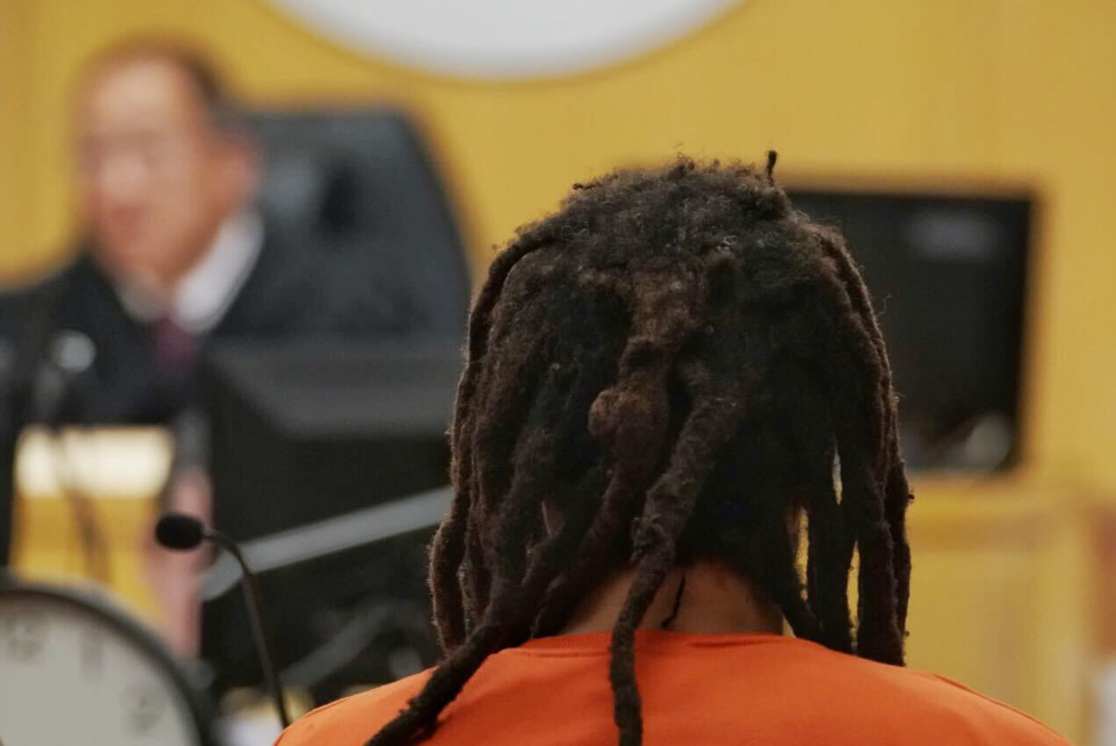 The back of a man in an orange shirt looks in the distance in a courtroom setting.