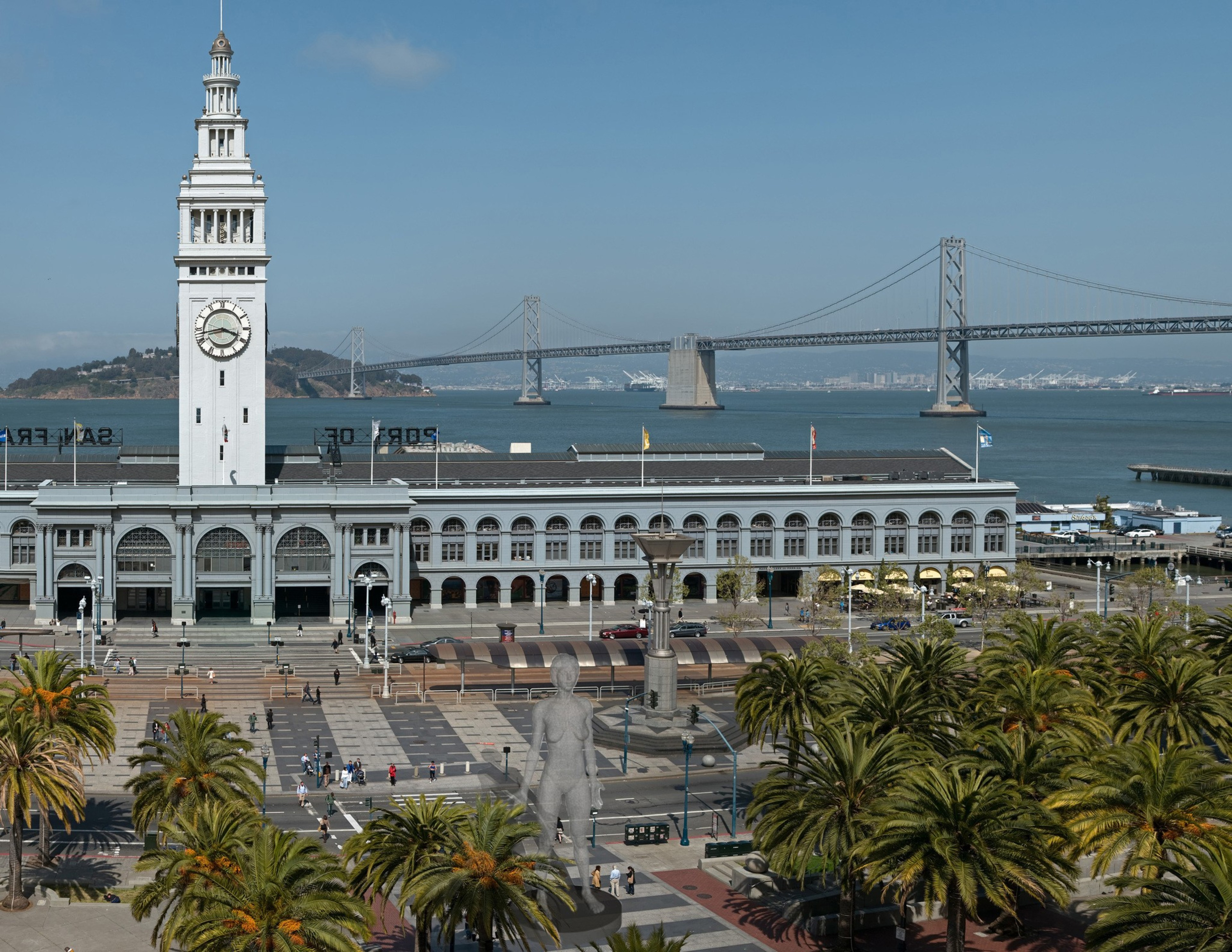 The image shows the San Francisco Ferry Building with a clock tower. The Bay Bridge is in the background, and palm trees line a plaza in the foreground.