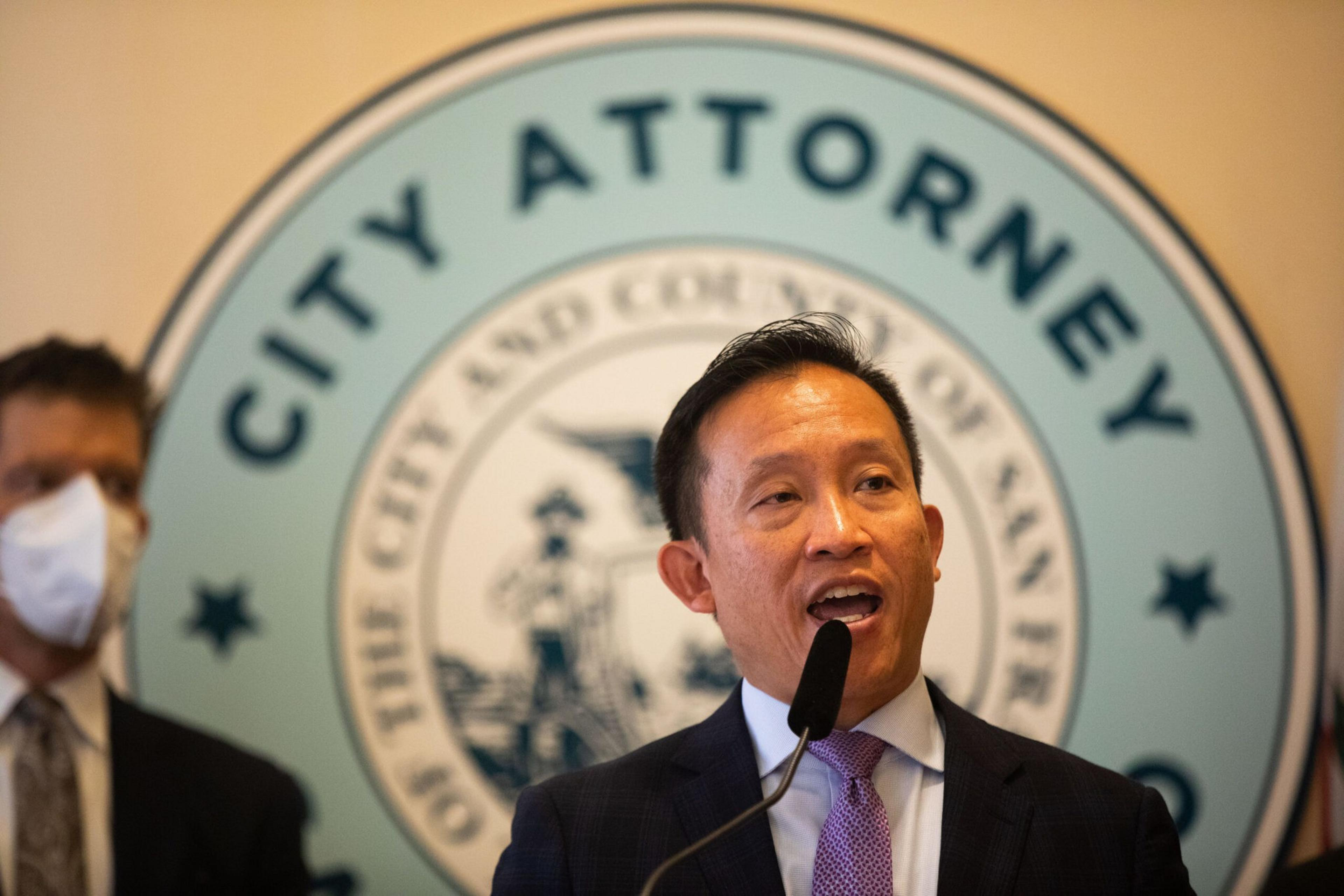 A man is speaking into a microphone with a "CITY ATTORNEY" seal in the background and another person partially visible on the left.