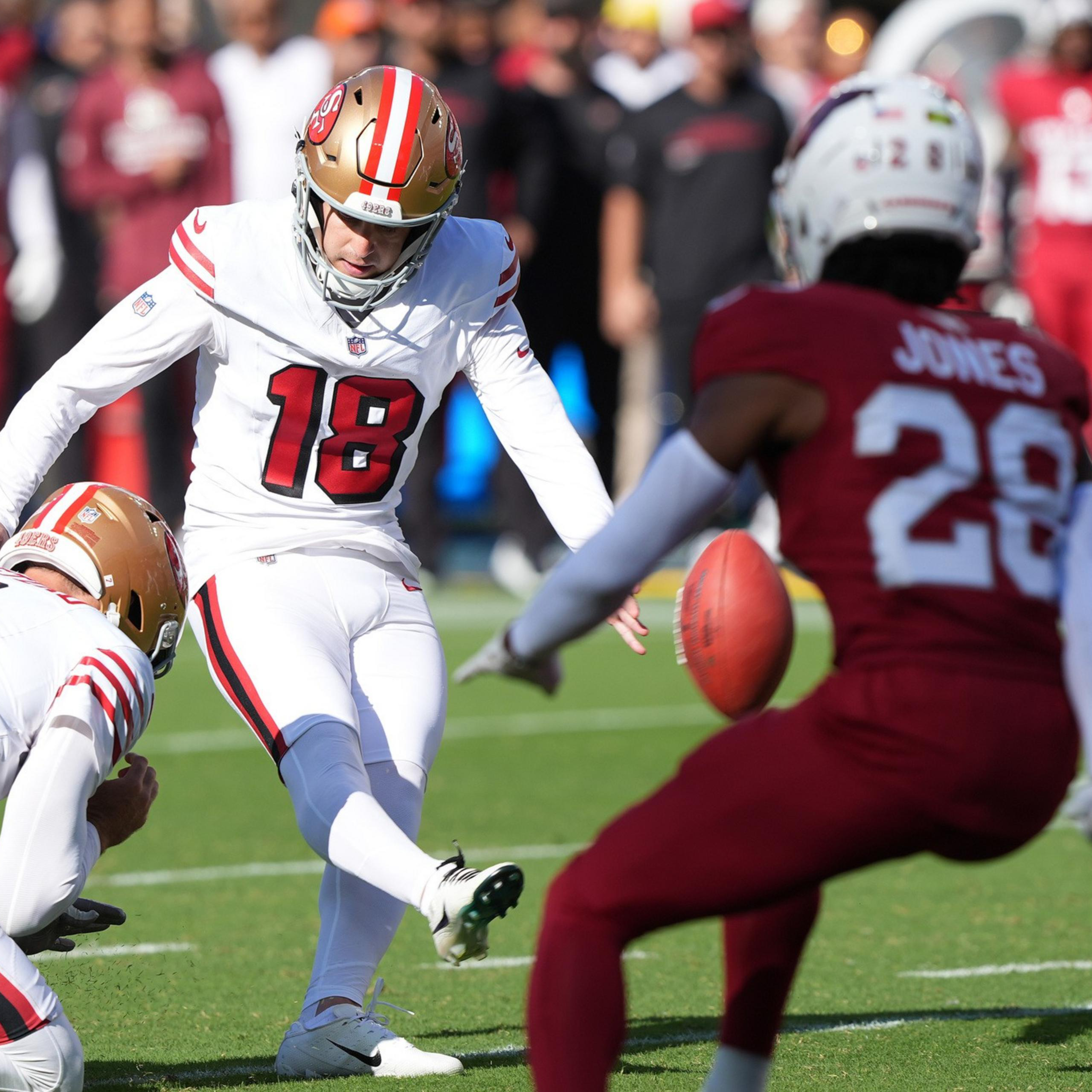A football player in white uniform #18 kicks the ball held by teammate #7, while a red-clad opponent #28 approaches on green field.