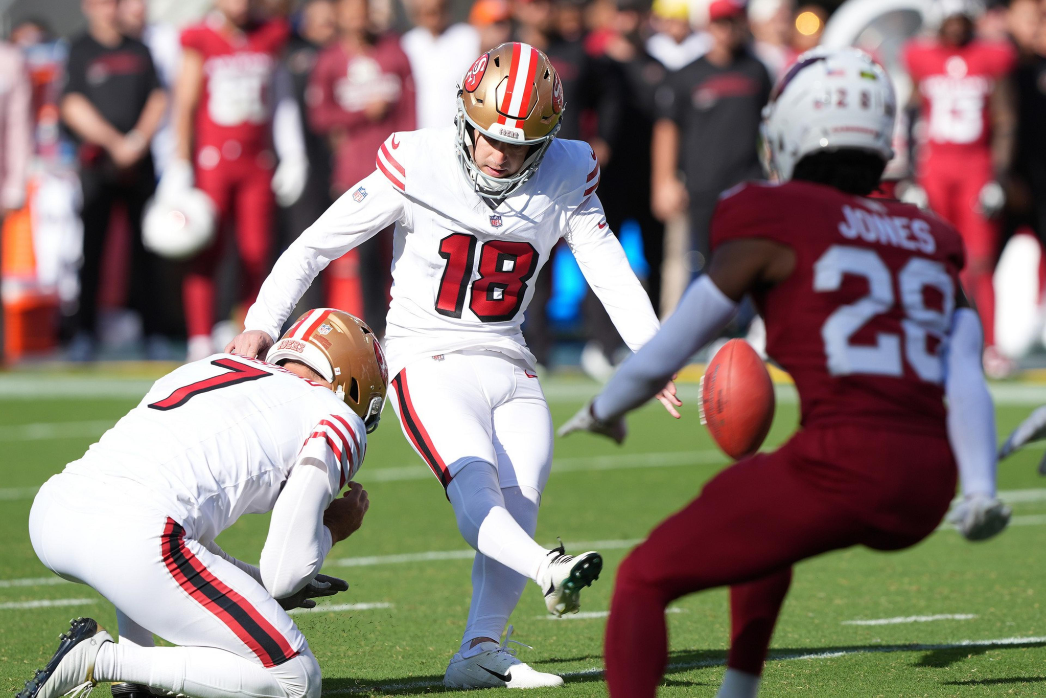 A football player in white uniform #18 kicks the ball held by teammate #7, while a red-clad opponent #28 approaches on green field.