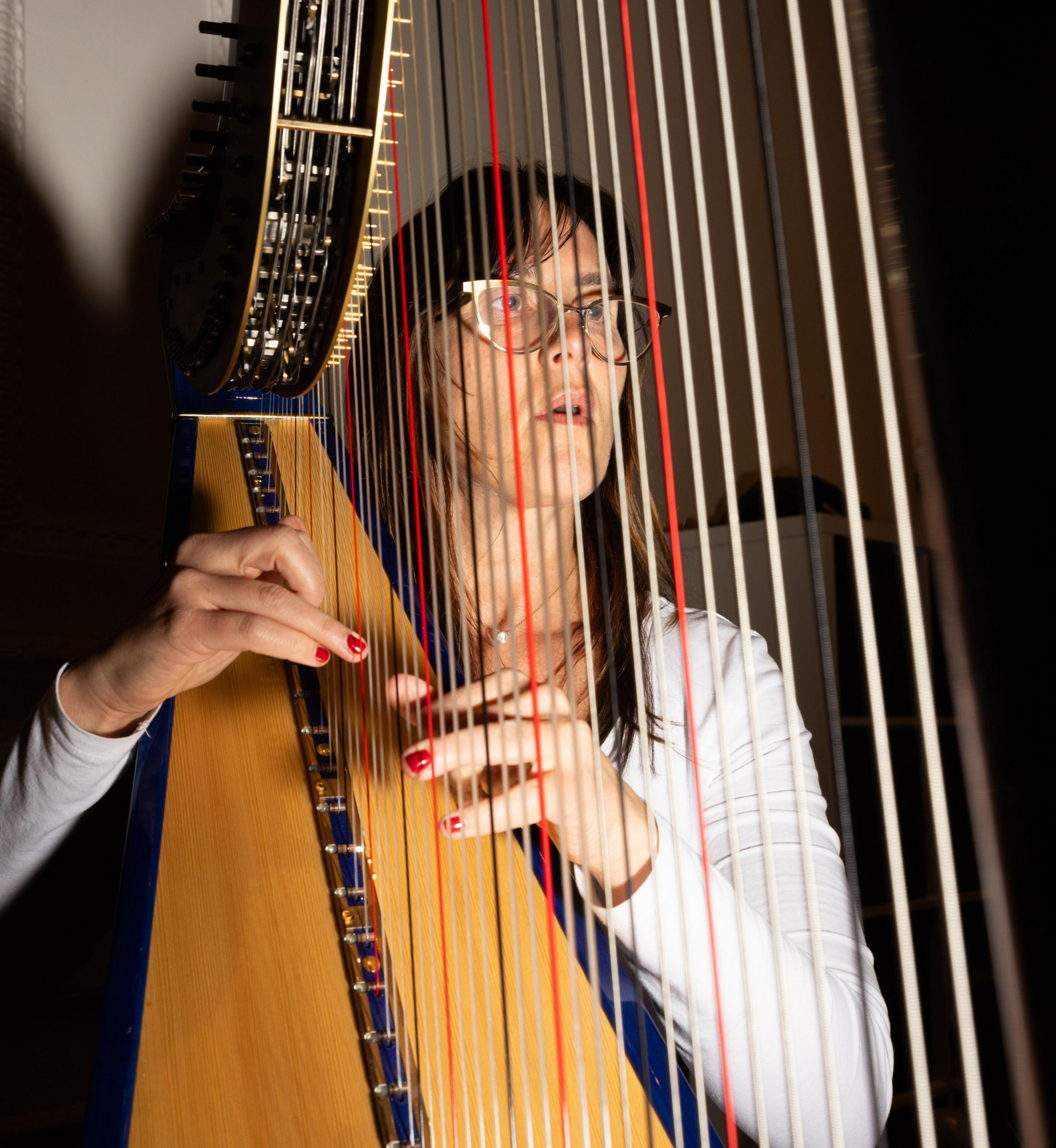 A person with glasses and red nail polish is playing a large harp, focusing intently on the instrument. The harp strings partially obscure their face.