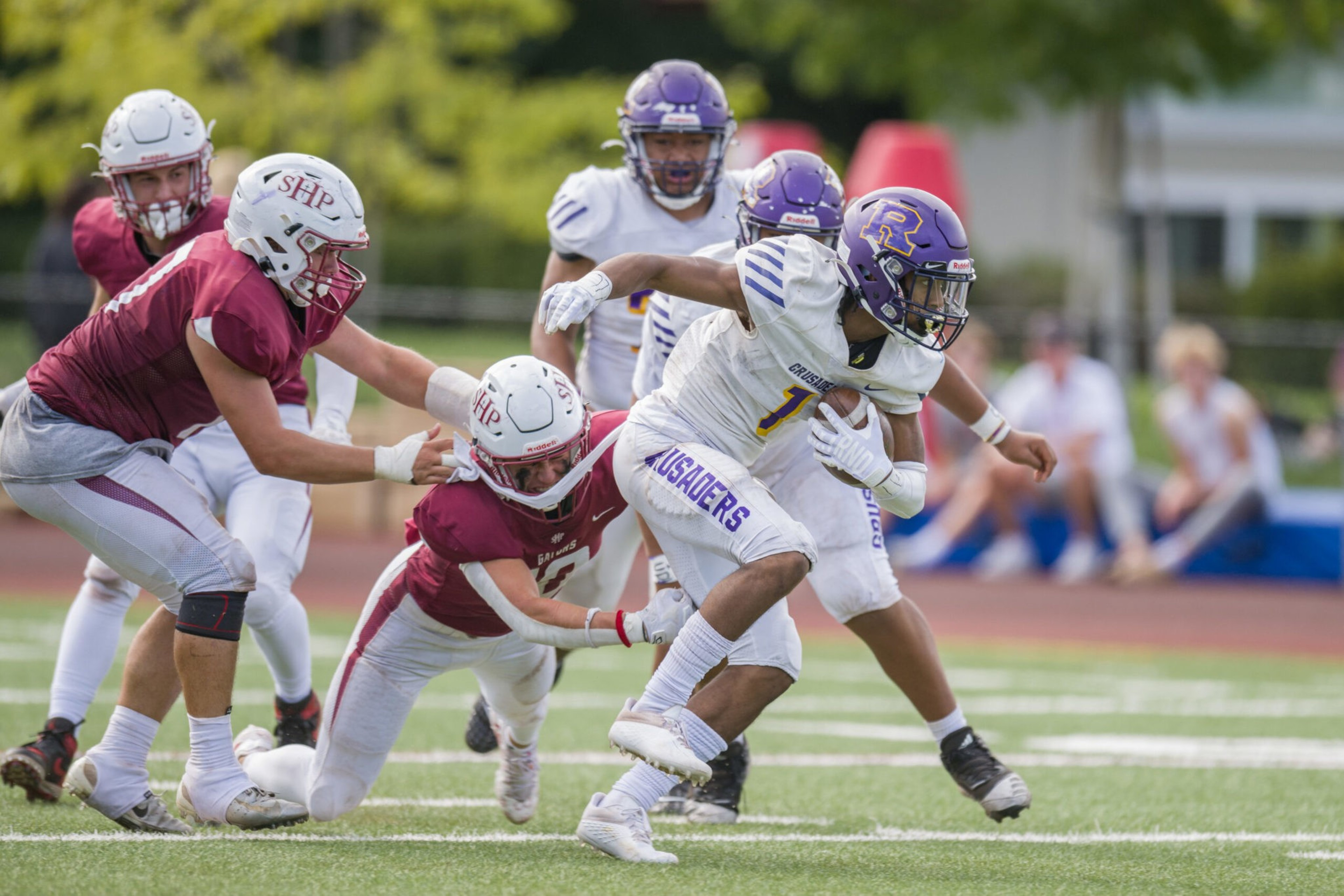 A football player in white and purple runs with the ball while two players in maroon and white try to tackle him on a green field.