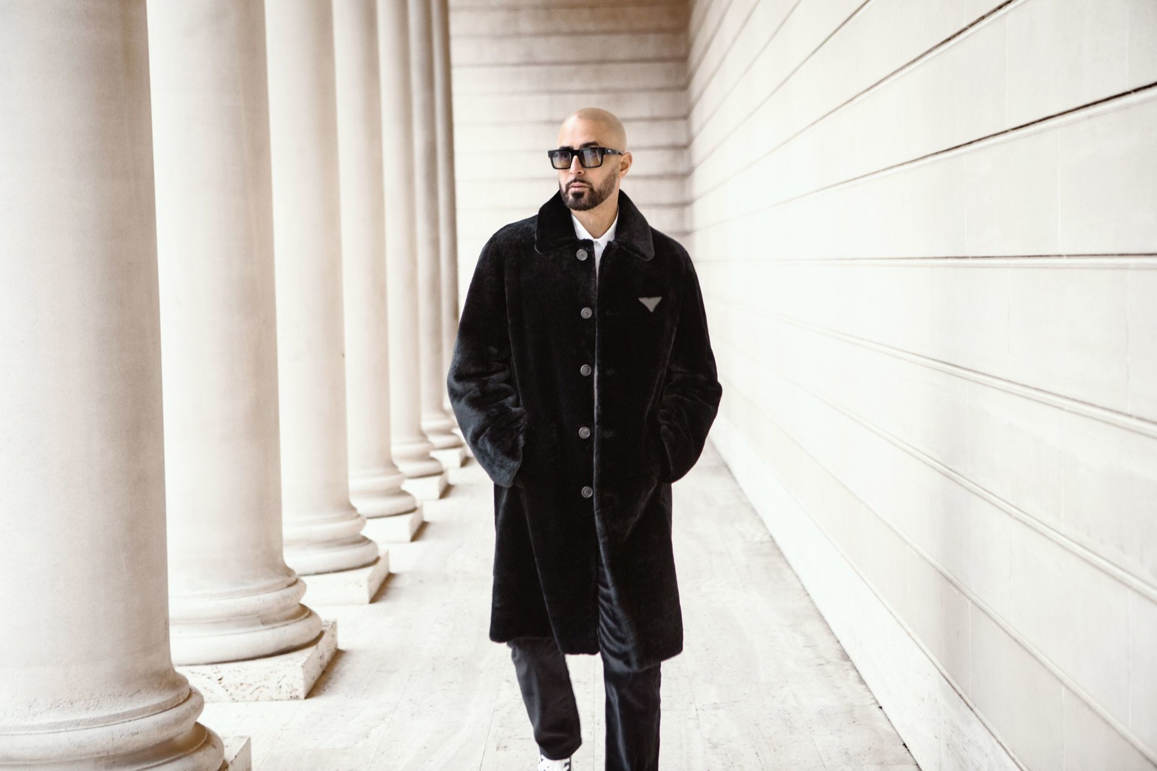 A person in a long black coat and sunglasses walks confidently through a corridor lined with tall stone columns, exuding an air of sophistication.