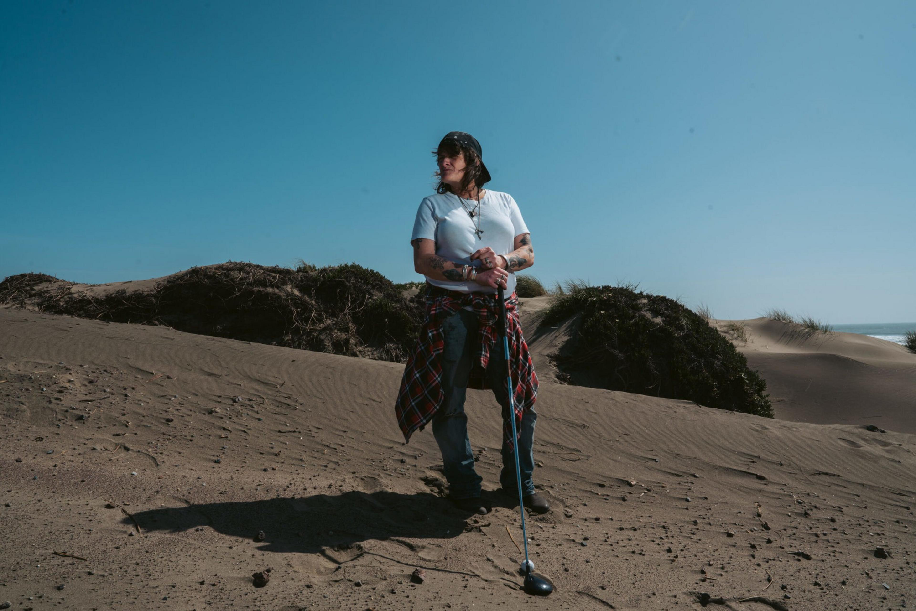 A person stands on sand dunes under a clear blue sky, holding a golf club, wearing a white shirt, jeans, and a plaid shirt tied around their waist.