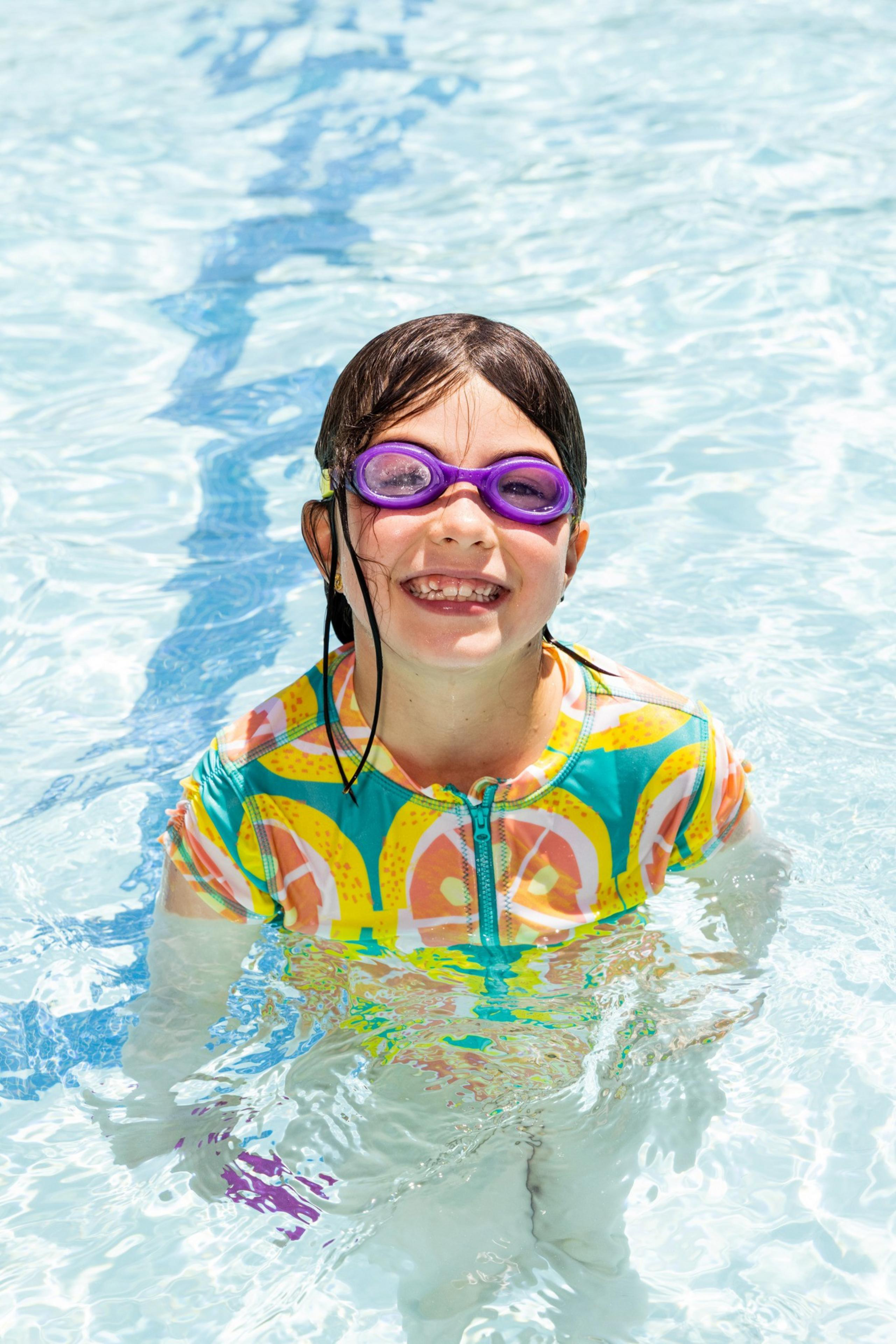 A child wearing purple swim goggles and a colorful swimsuit with fruit designs is happily standing in a pool, smiling widely with their hair wet.