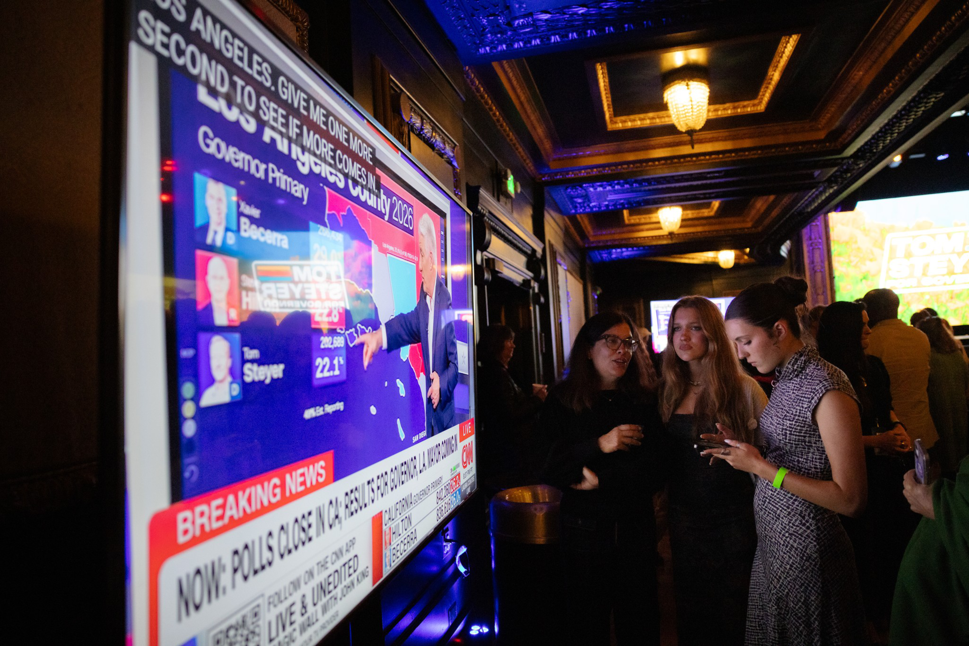 People in a dimly lit room watch a large TV screen displaying California 2026 governor primary election results and breaking news coverage.