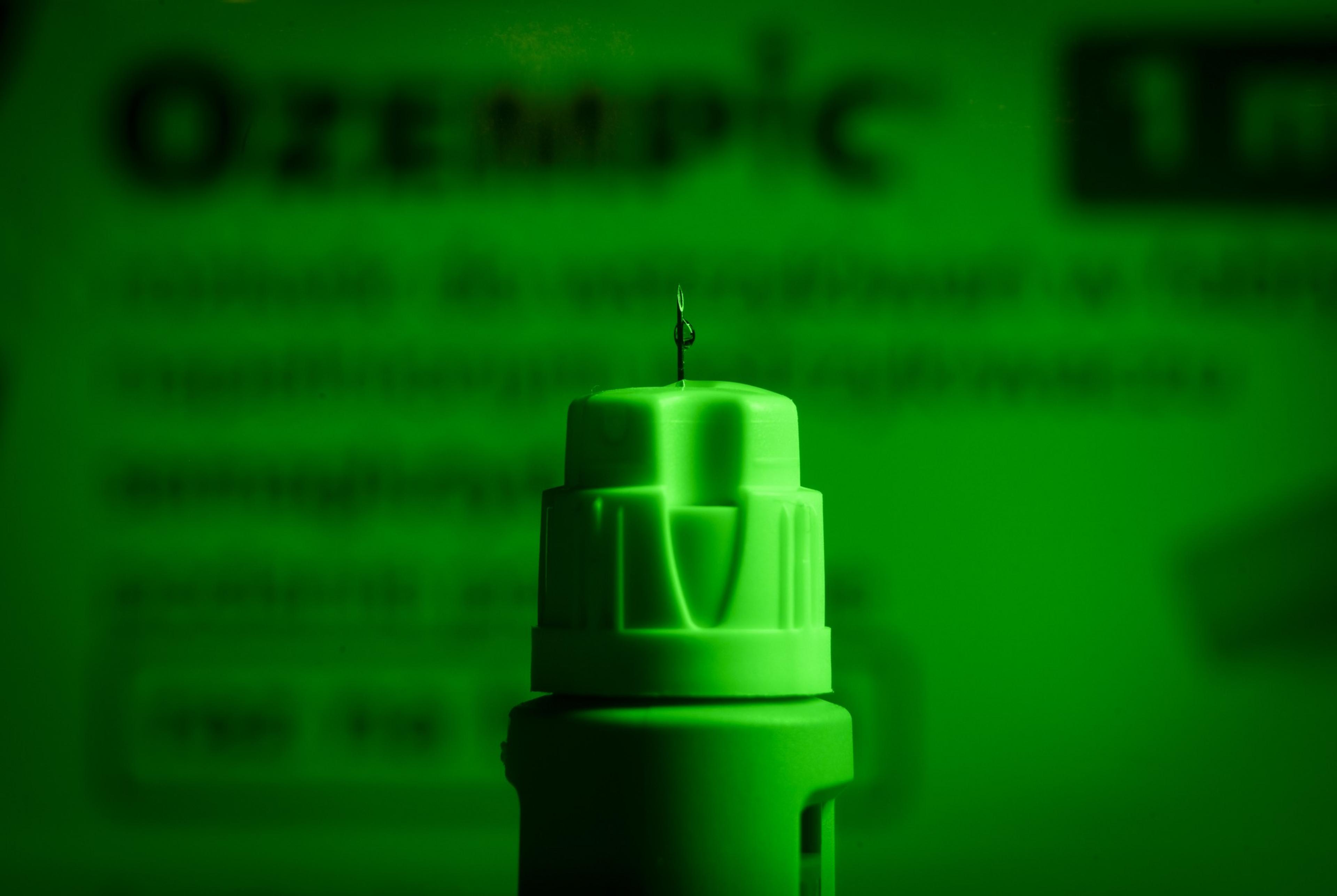 A close-up of a green injection pen with a droplet on its needle, set against a faintly visible blurred text backdrop.