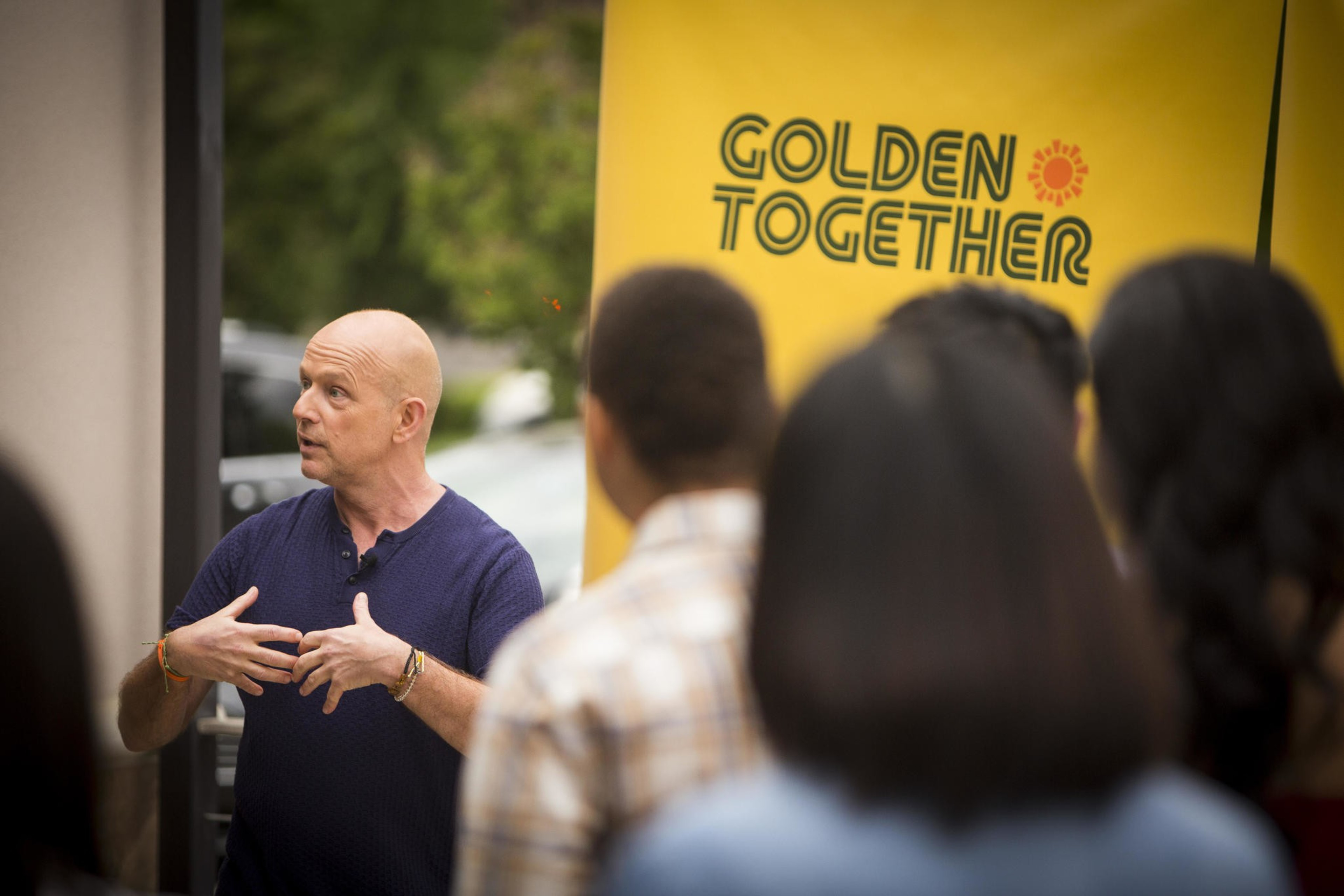 A bald man in a blue shirt speaks animatedly to a group. Behind him is a yellow banner with &quot;Golden Together&quot; and a sun graphic. People listen attentively.