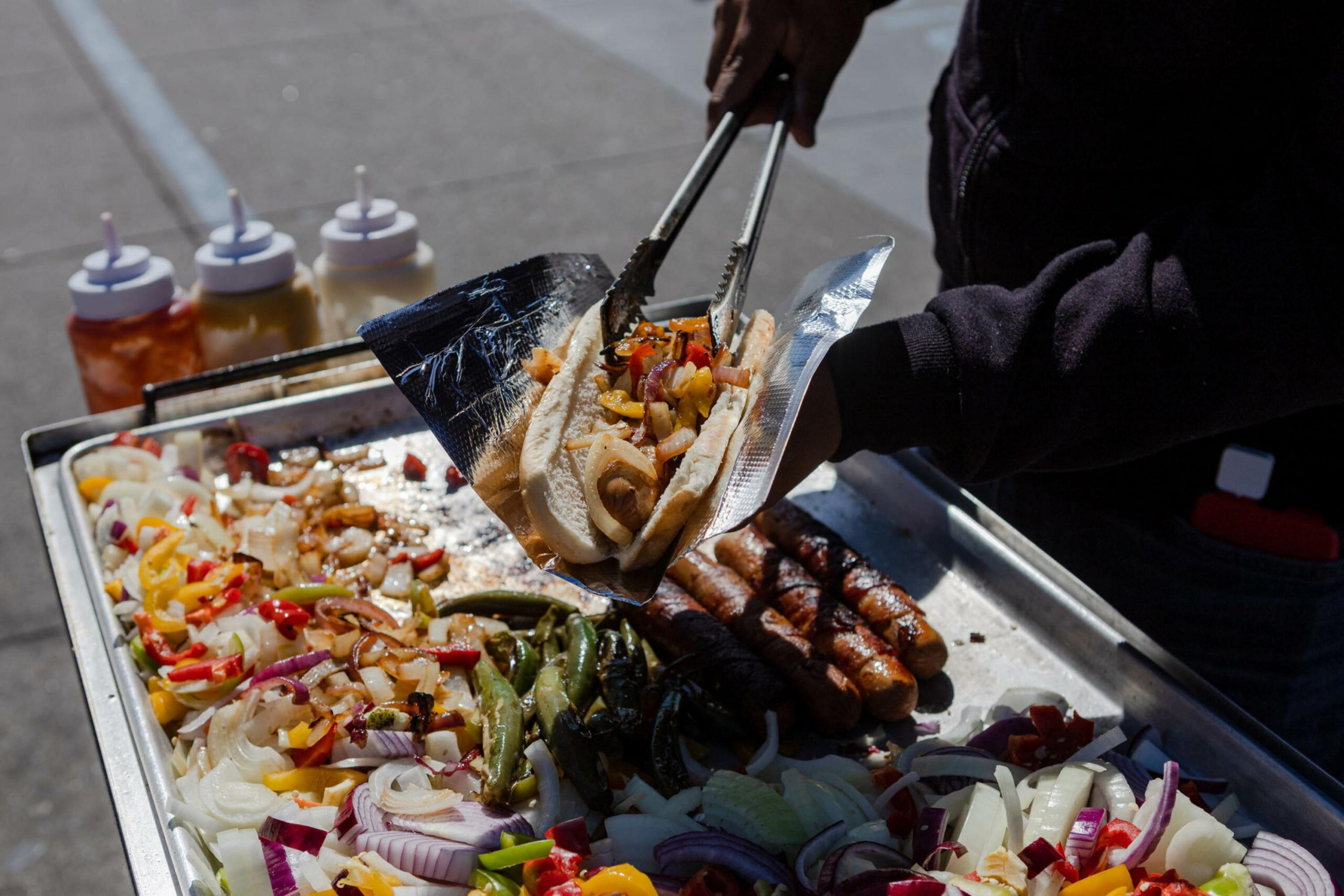 A person uses tongs to place grilled sausages and mixed peppers with onions into a bun on a food cart with condiments nearby.