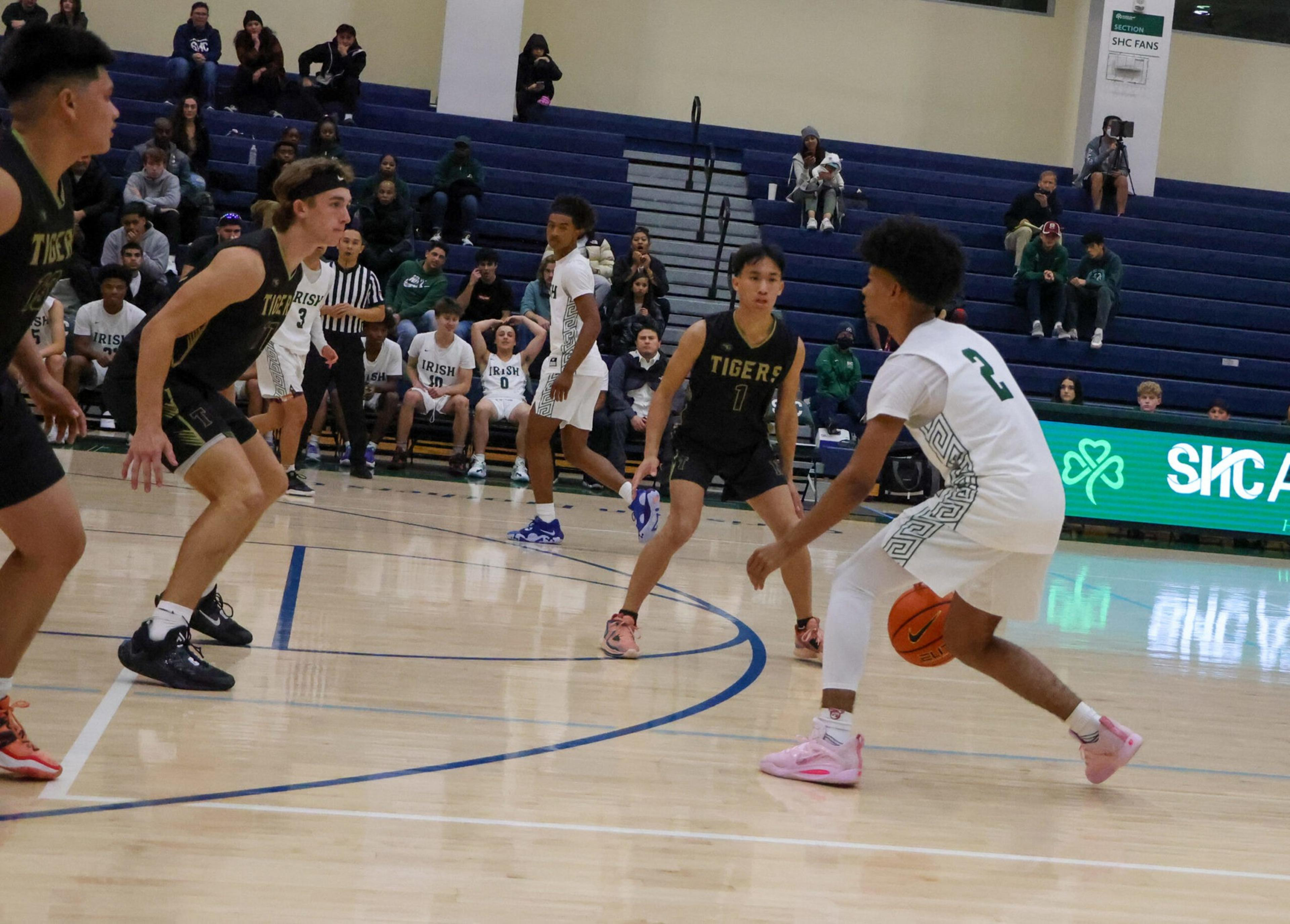 A basketball player in white dribbles the ball while three defenders in black jerseys prepare to block him on the court.