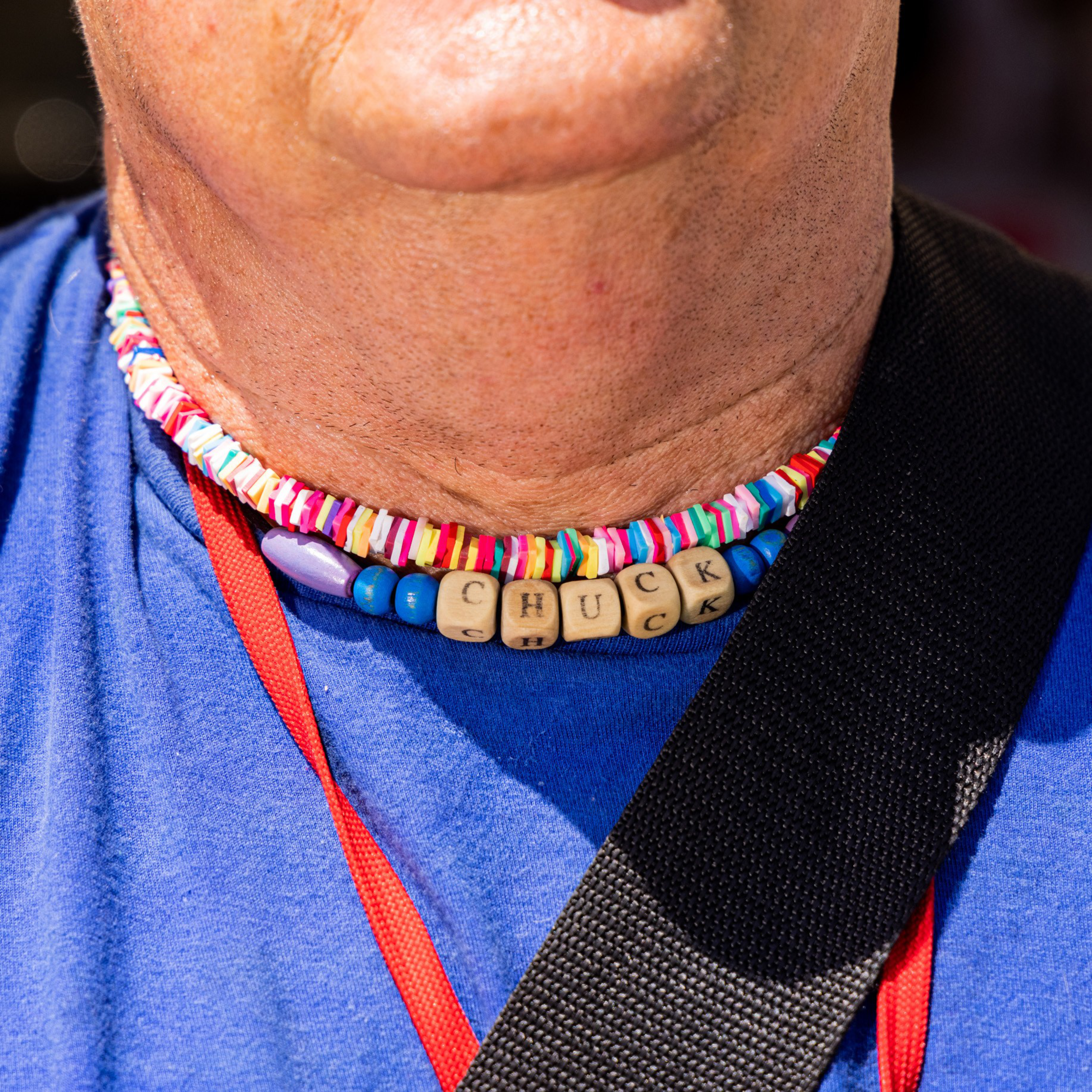 The image shows a person wearing colorful necklaces, including one with wooden beads spelling &quot;CHUCK,&quot; and dressed in a blue shirt with red and black straps.