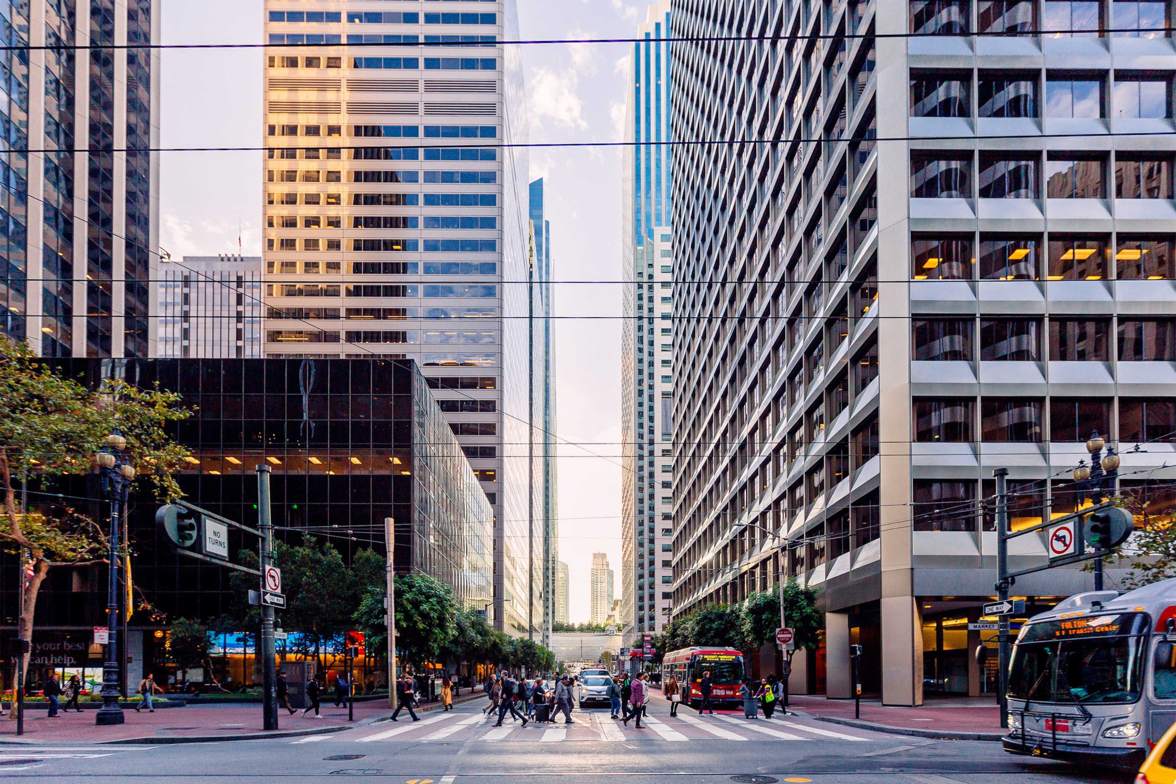 A city street lined with tall buildings, pedestrians crossing, a bus, and traffic lights at dusk.