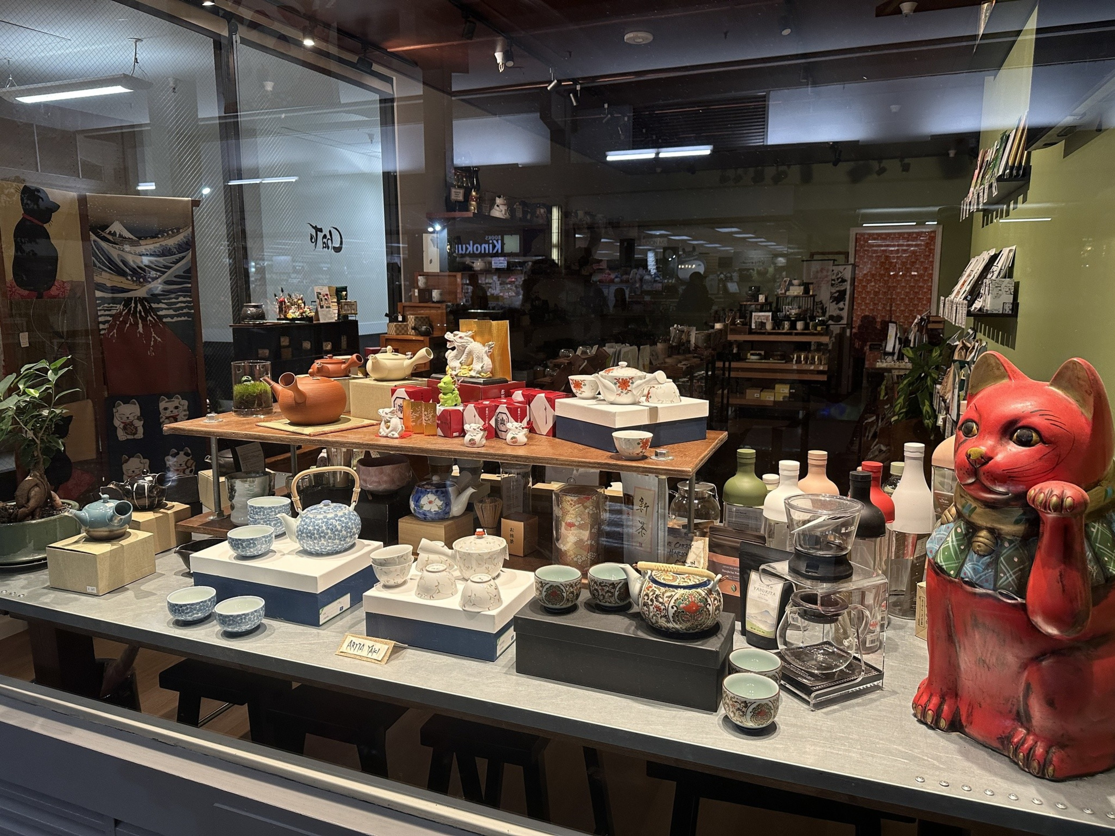 A shop window is filled with colorful, Japanese-style tea pots and a statue of a red cat. 