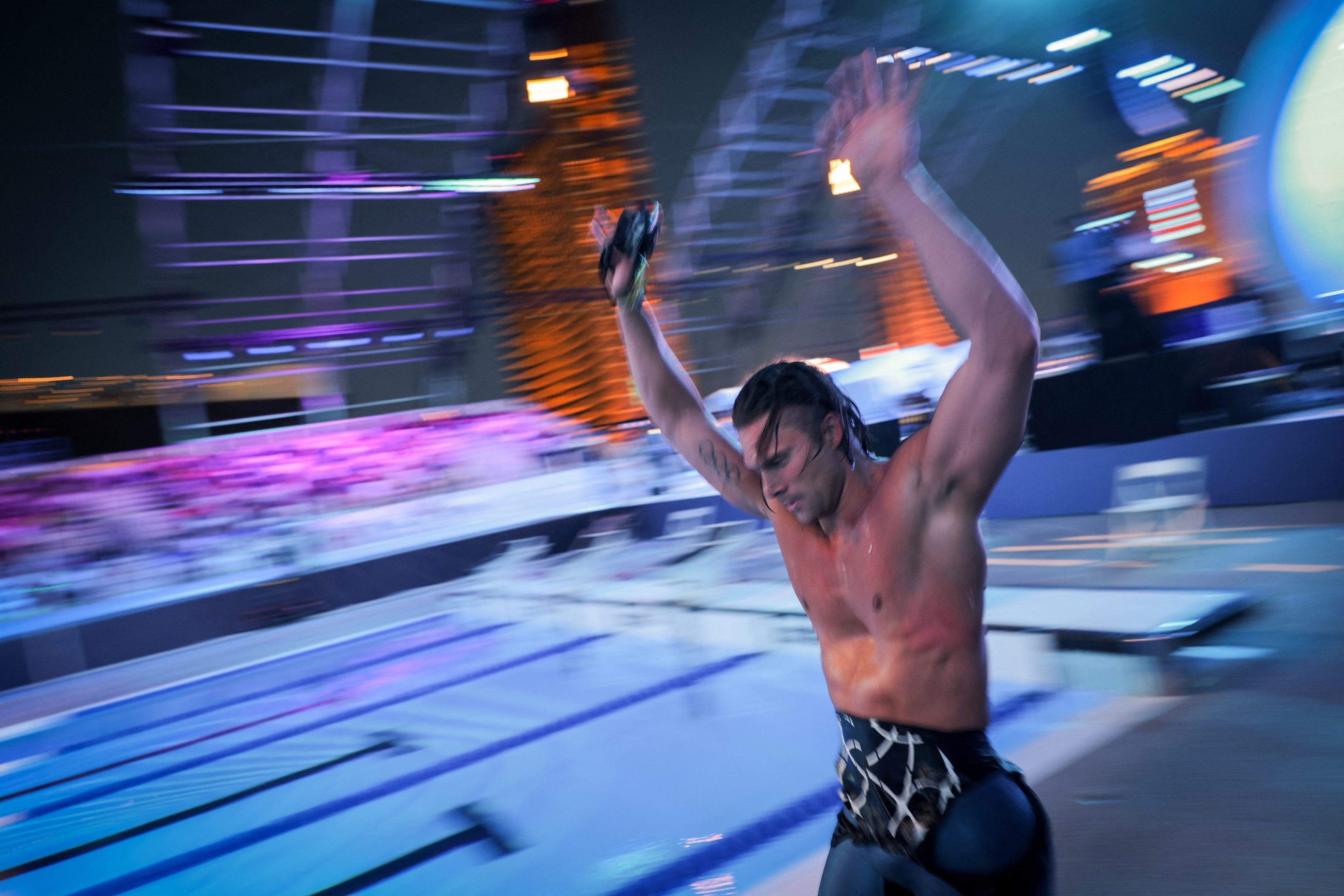 A shirtless man with wet hair raises his arms near a brightly lit swimming pool at night, appearing to celebrate or prepare to dive.