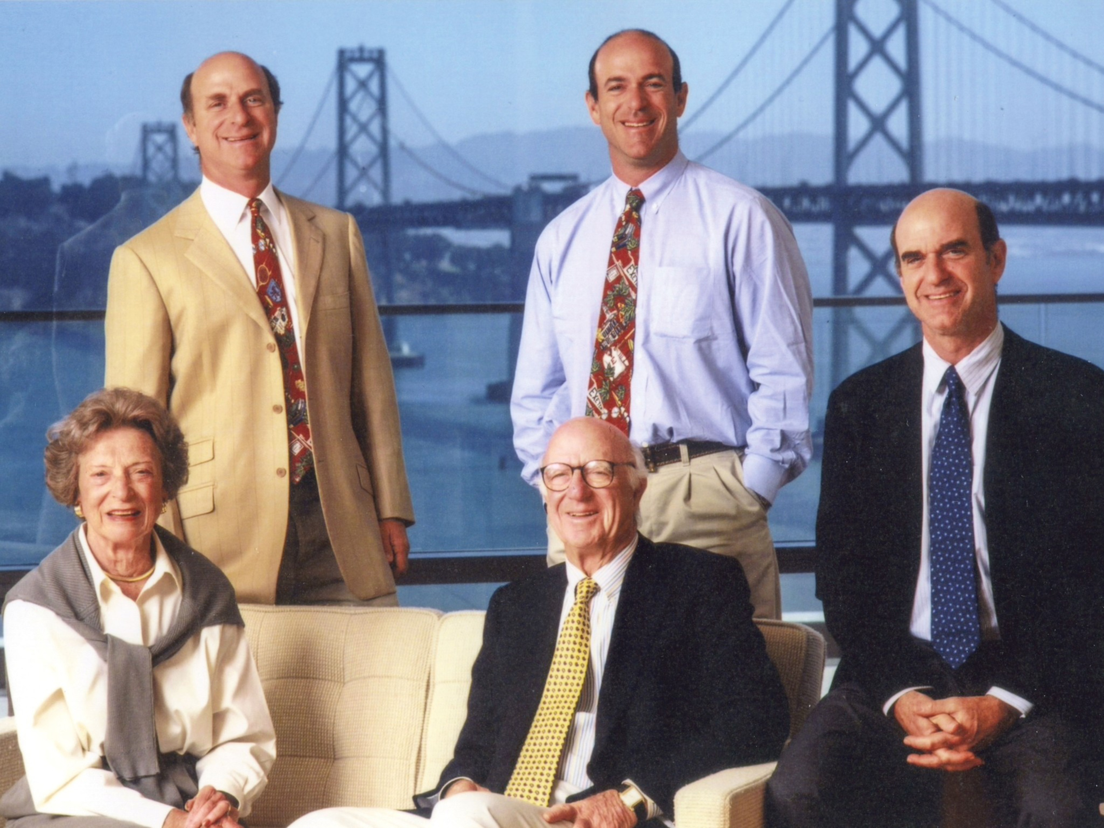 Five smiling people, three men standing and two seated, dressed in business attire, with a large suspension bridge and water in the background.
