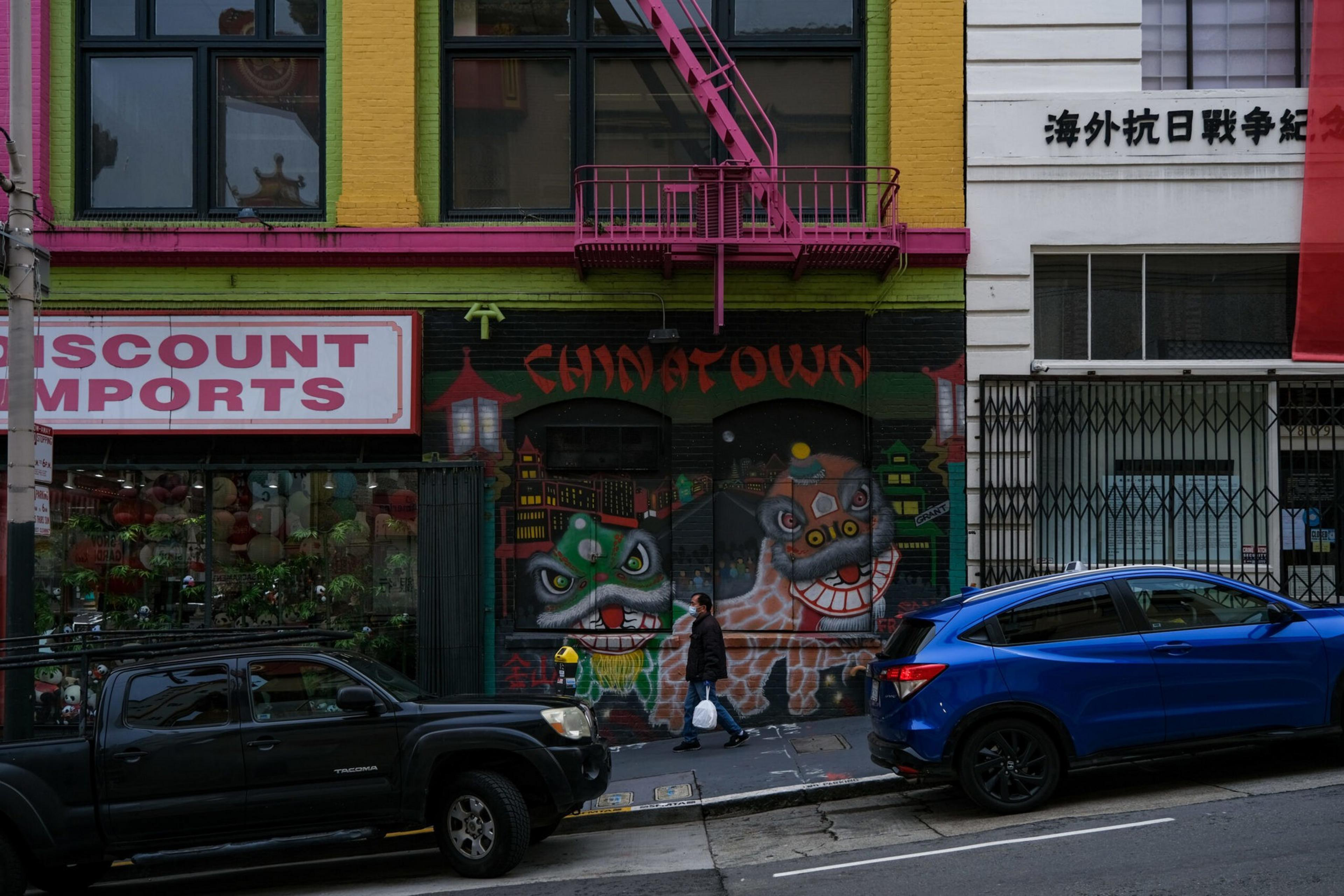 A person walks past a colorful Chinatown mural with two lion dance faces on a building wall between parked black and blue cars.