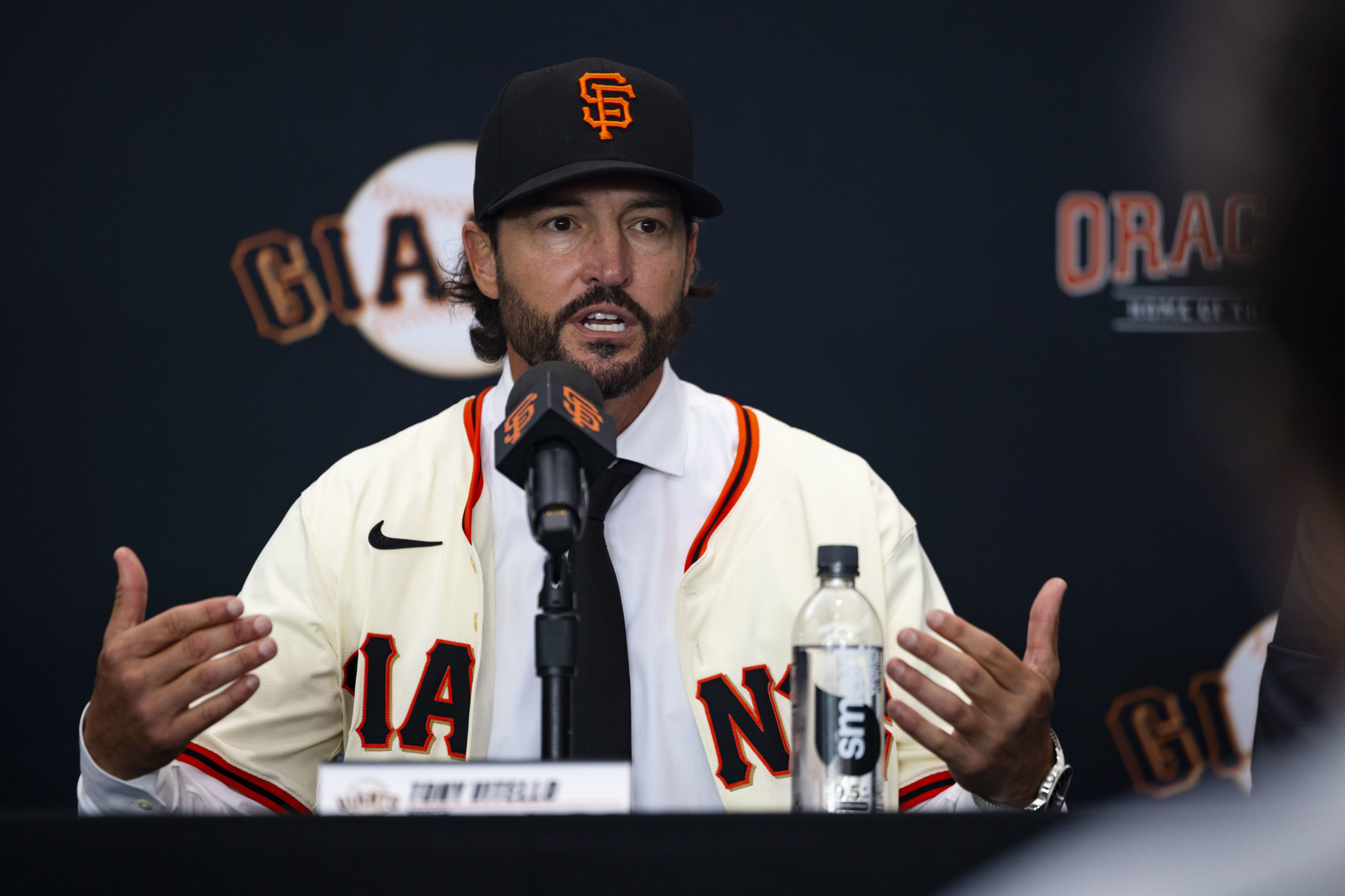 A man wearing a San Francisco Giants baseball cap and jersey speaks into a microphone at a press conference with a water bottle in front of him.
