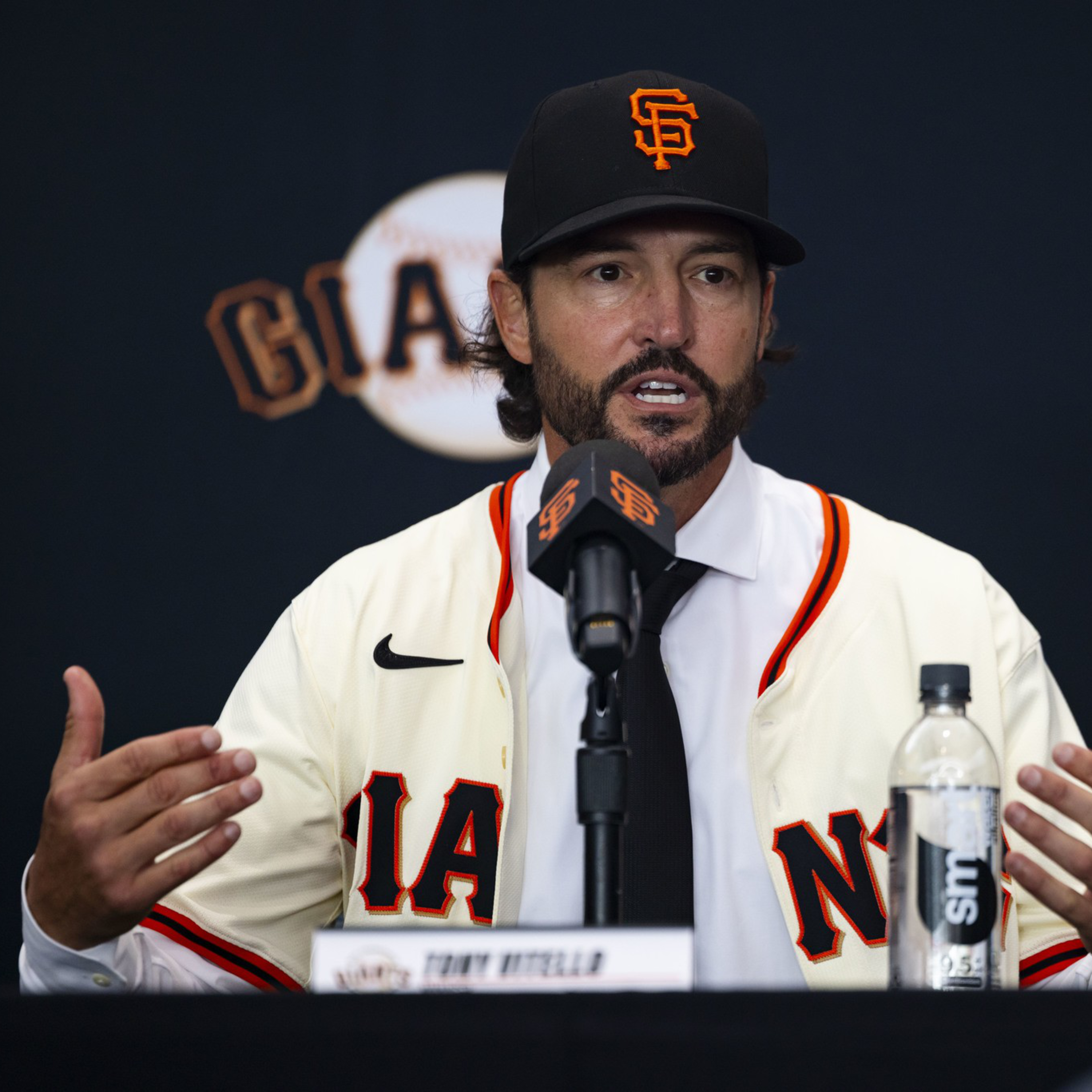 A man wearing a San Francisco Giants baseball cap and jersey speaks into a microphone at a press conference with a water bottle in front of him.