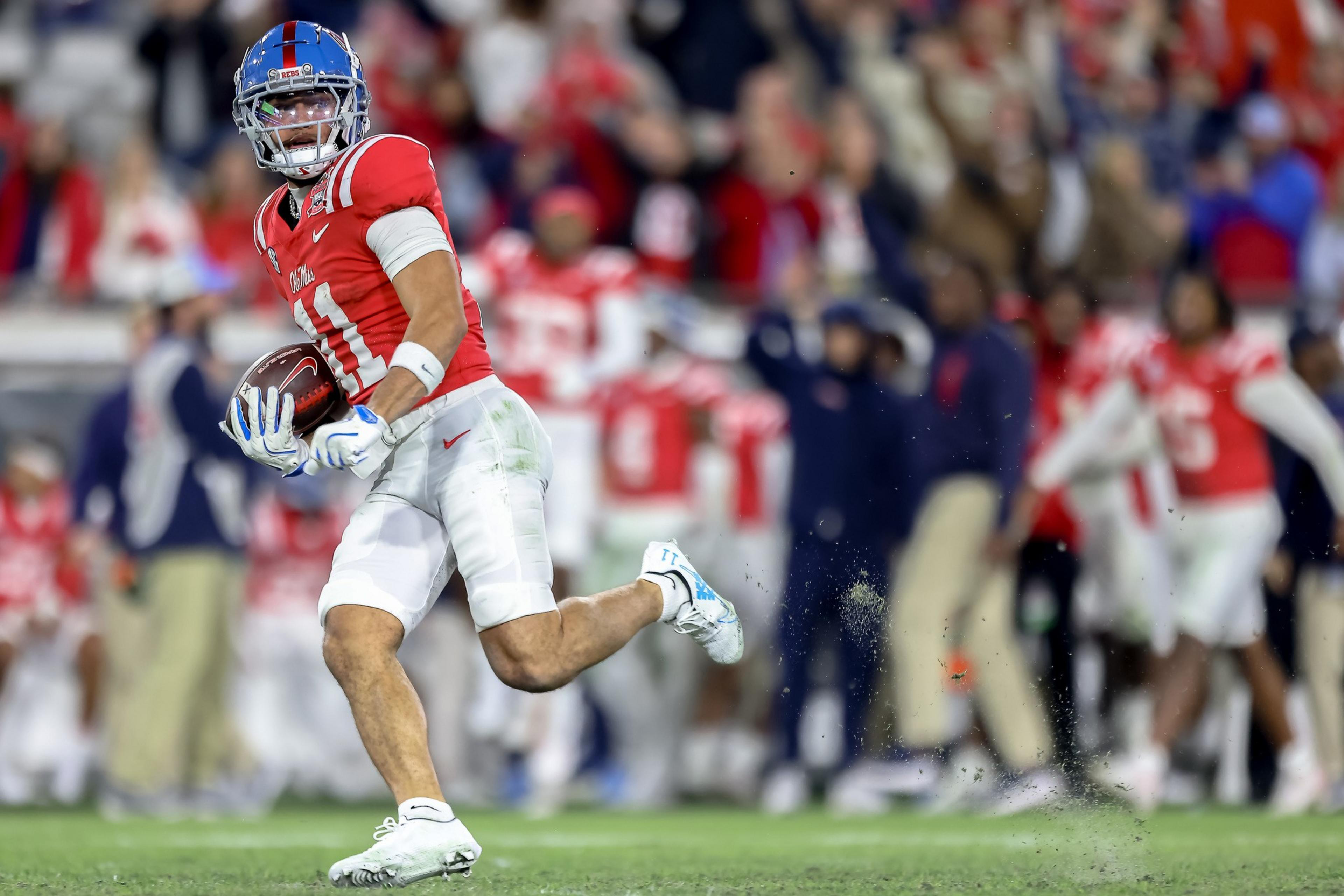 A football player in a red jersey and helmet runs on a grassy field, holding the ball tightly. The background shows a blurred crowd in similar attire.