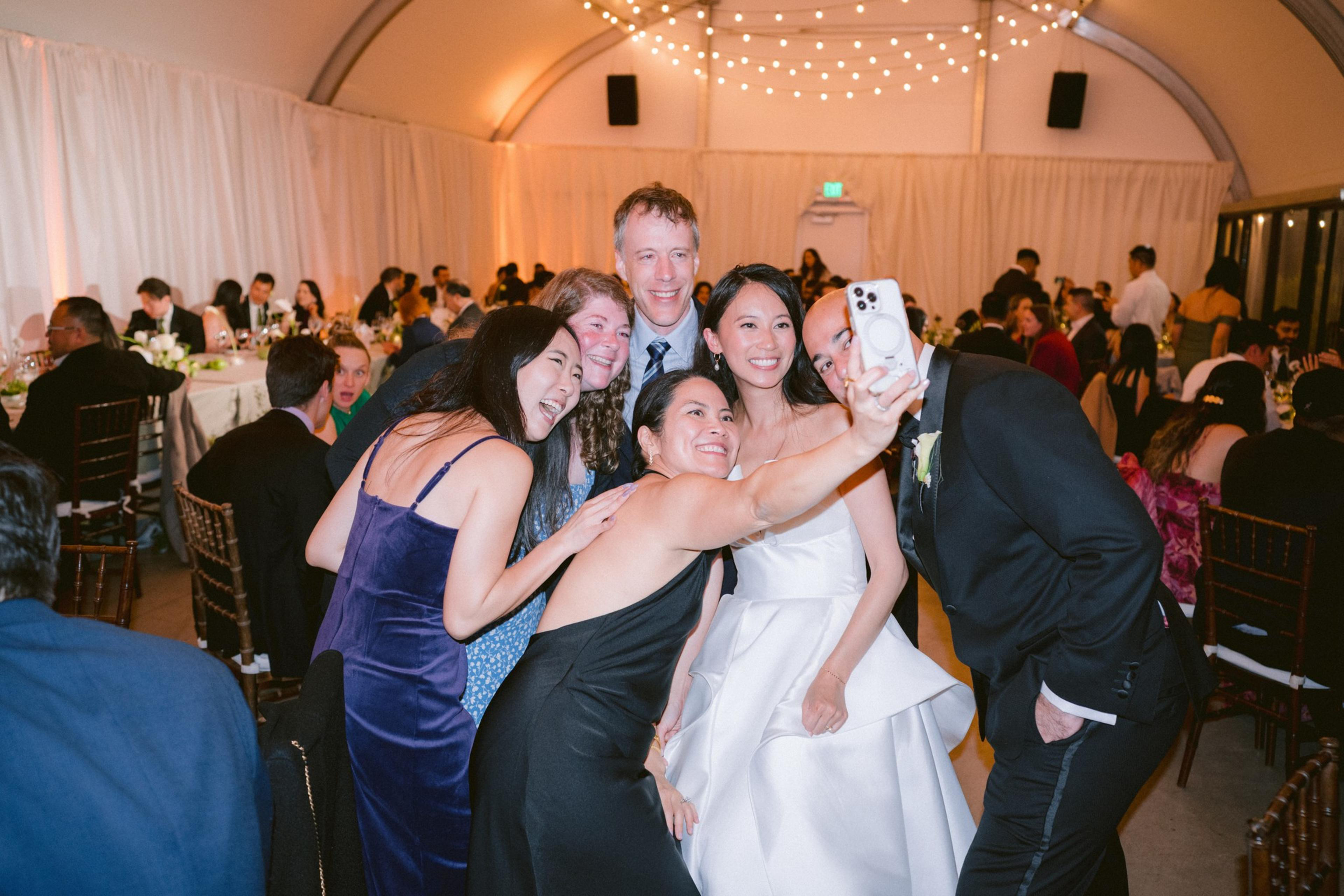 Five people, including a bride in a white dress, smile closely as they pose for a selfie at a decorated indoor wedding reception.