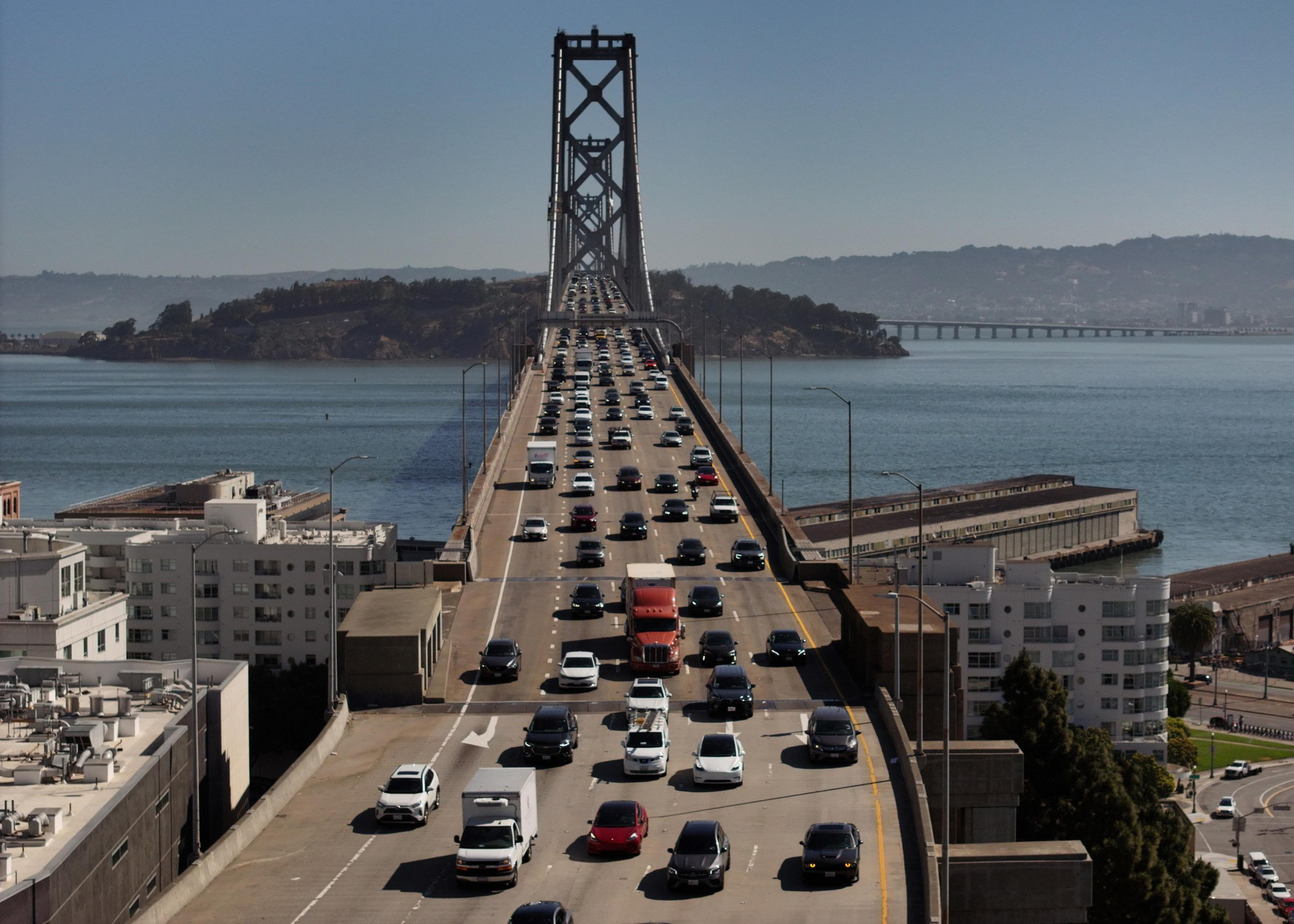 The image shows a busy suspension bridge with multiple lanes of traffic spanning across a body of water, connecting to a distant, hilly landscape.