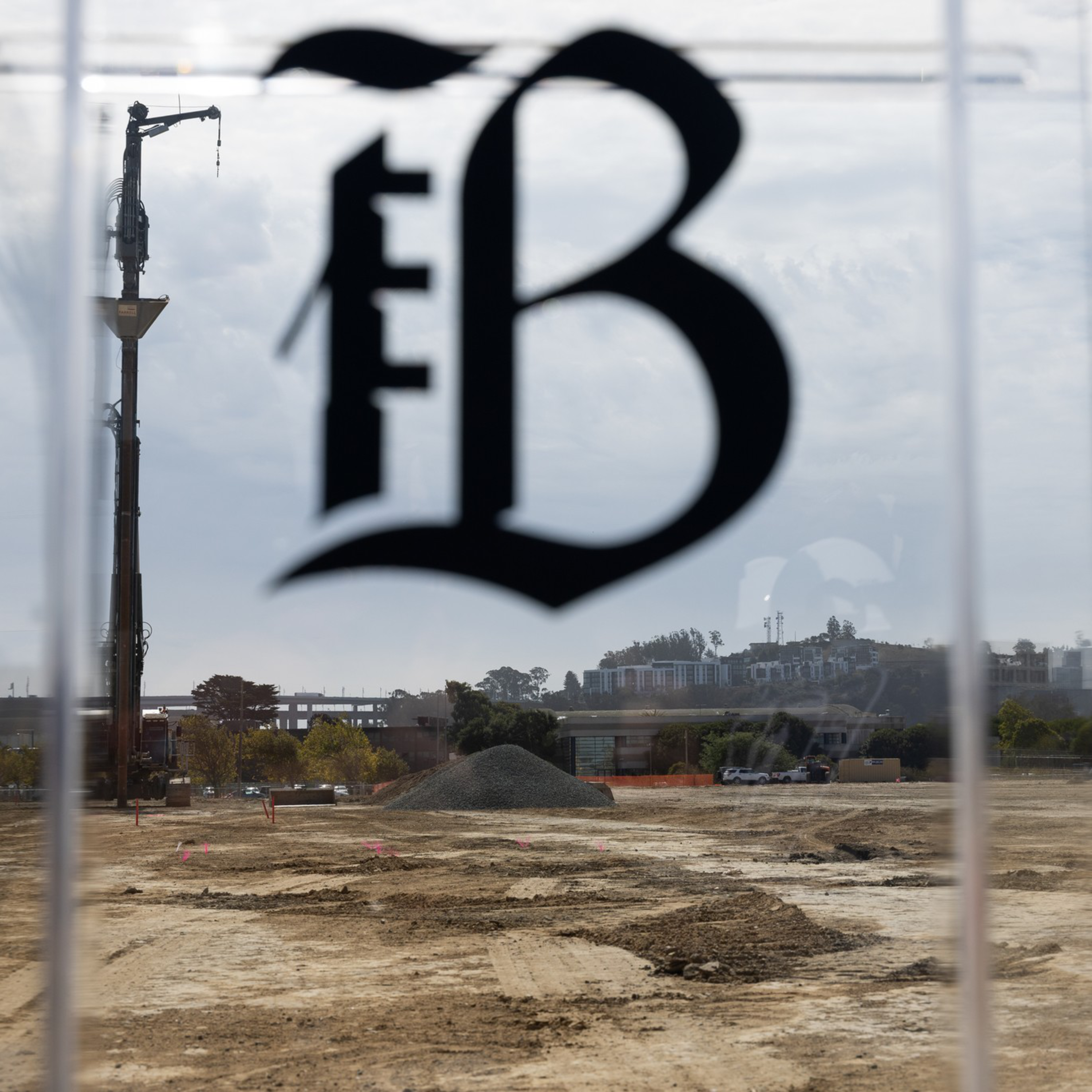A large construction site with dirt ground, a pile of gravel, cranes, and construction equipment is visible through a transparent window marked with a stylized "B."