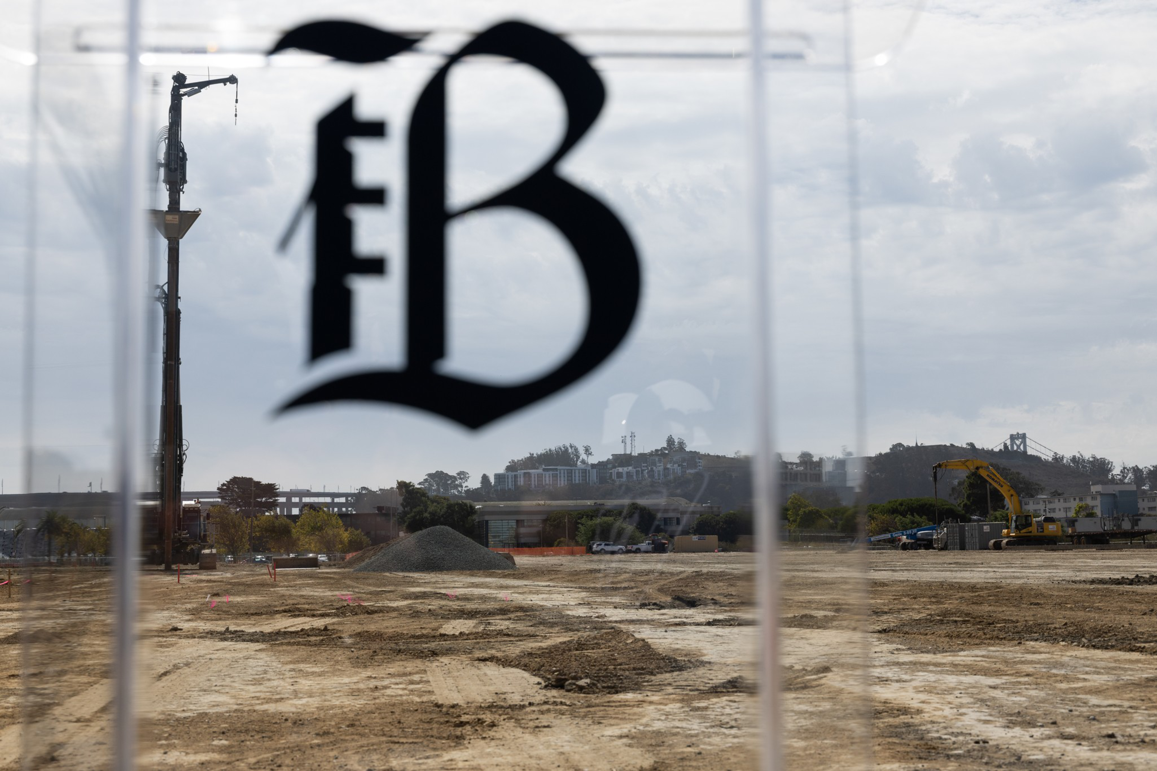 A large construction site with dirt ground, a pile of gravel, cranes, and construction equipment is visible through a transparent window marked with a stylized "B."