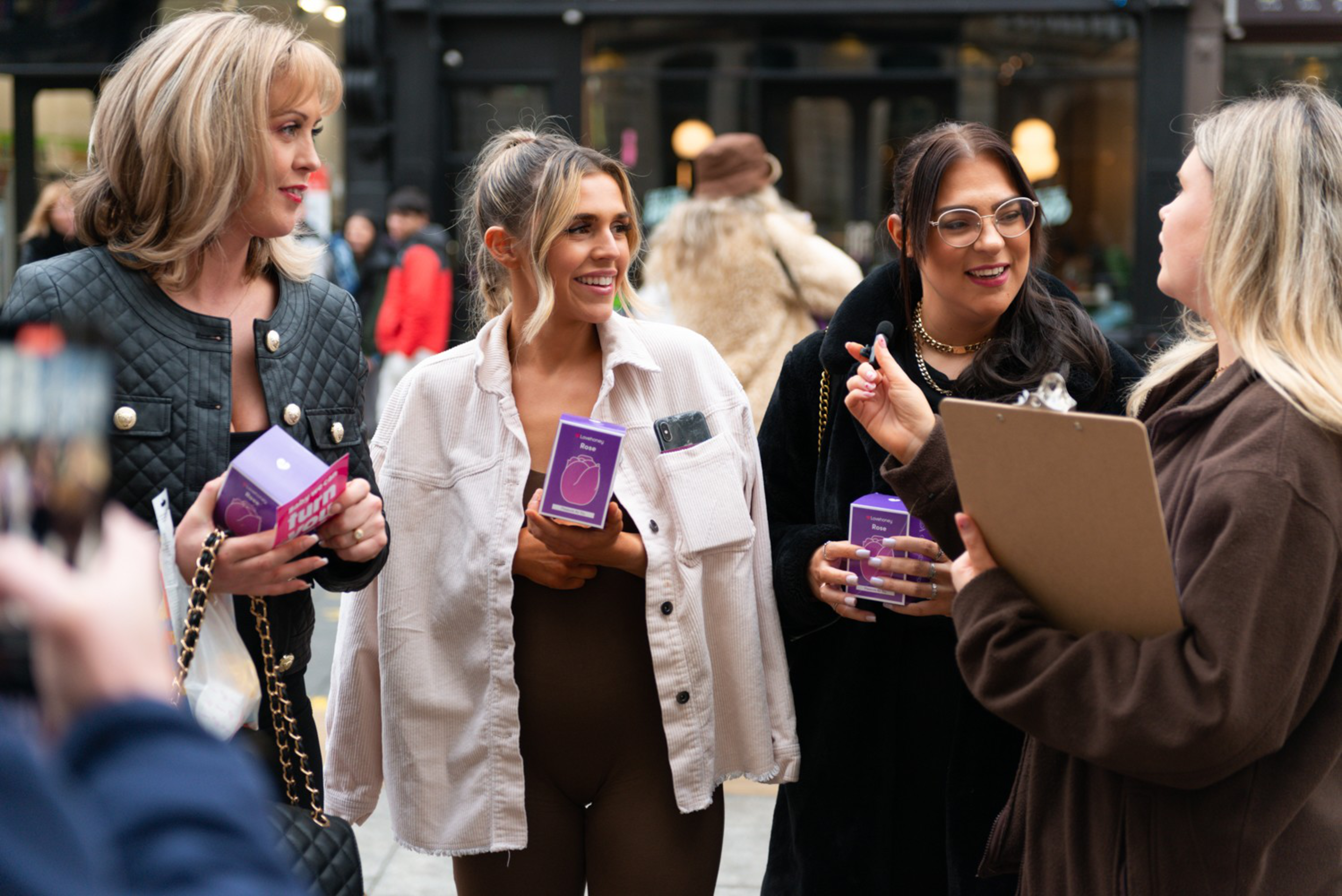 Four women are standing on a street, talking and holding purple boxes. One holds a clipboard and appears to be leading a conversation.