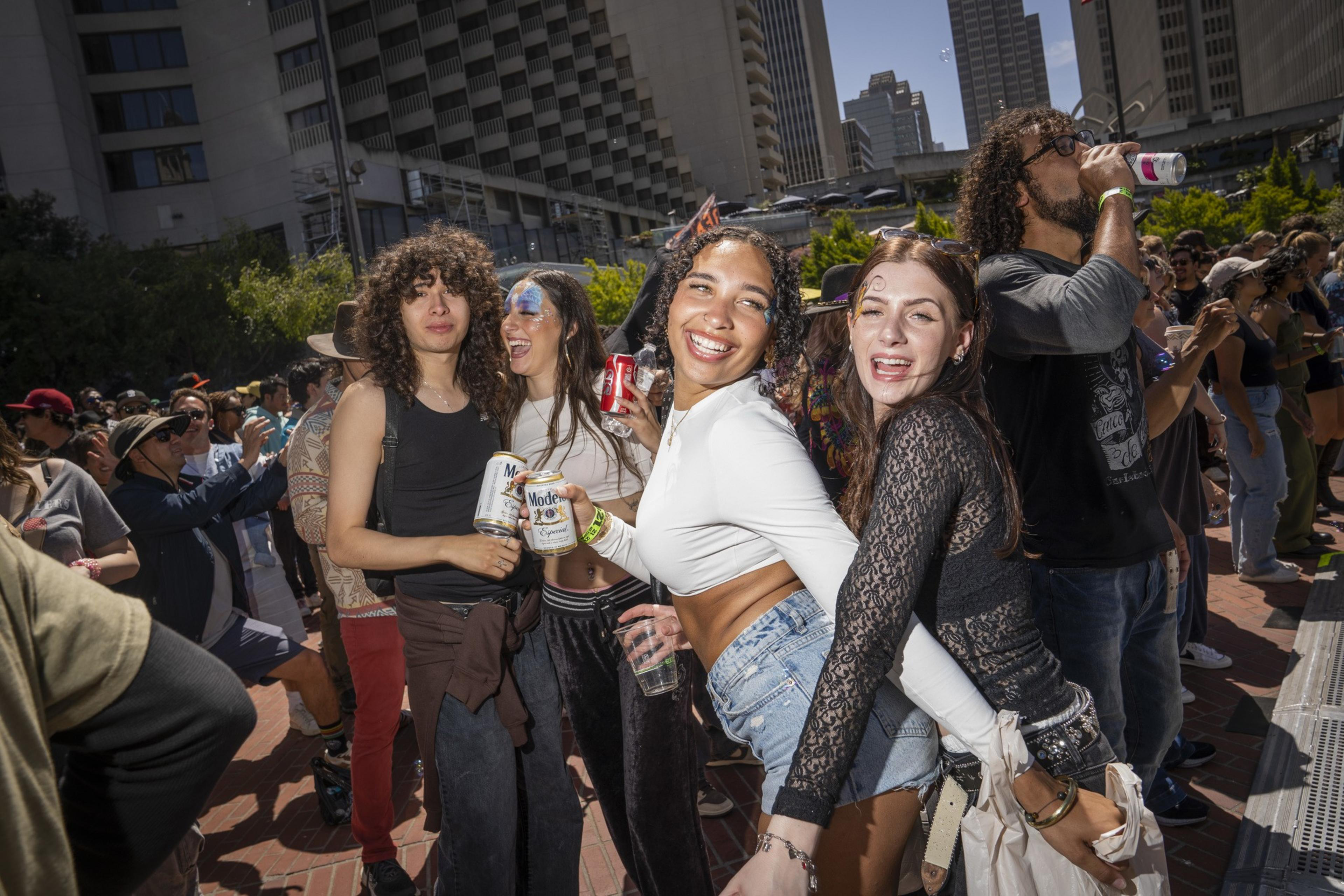 A joyful group is dancing outside at a lively event. They hold drinks and are surrounded by a crowd under clear blue skies, with buildings in the background.