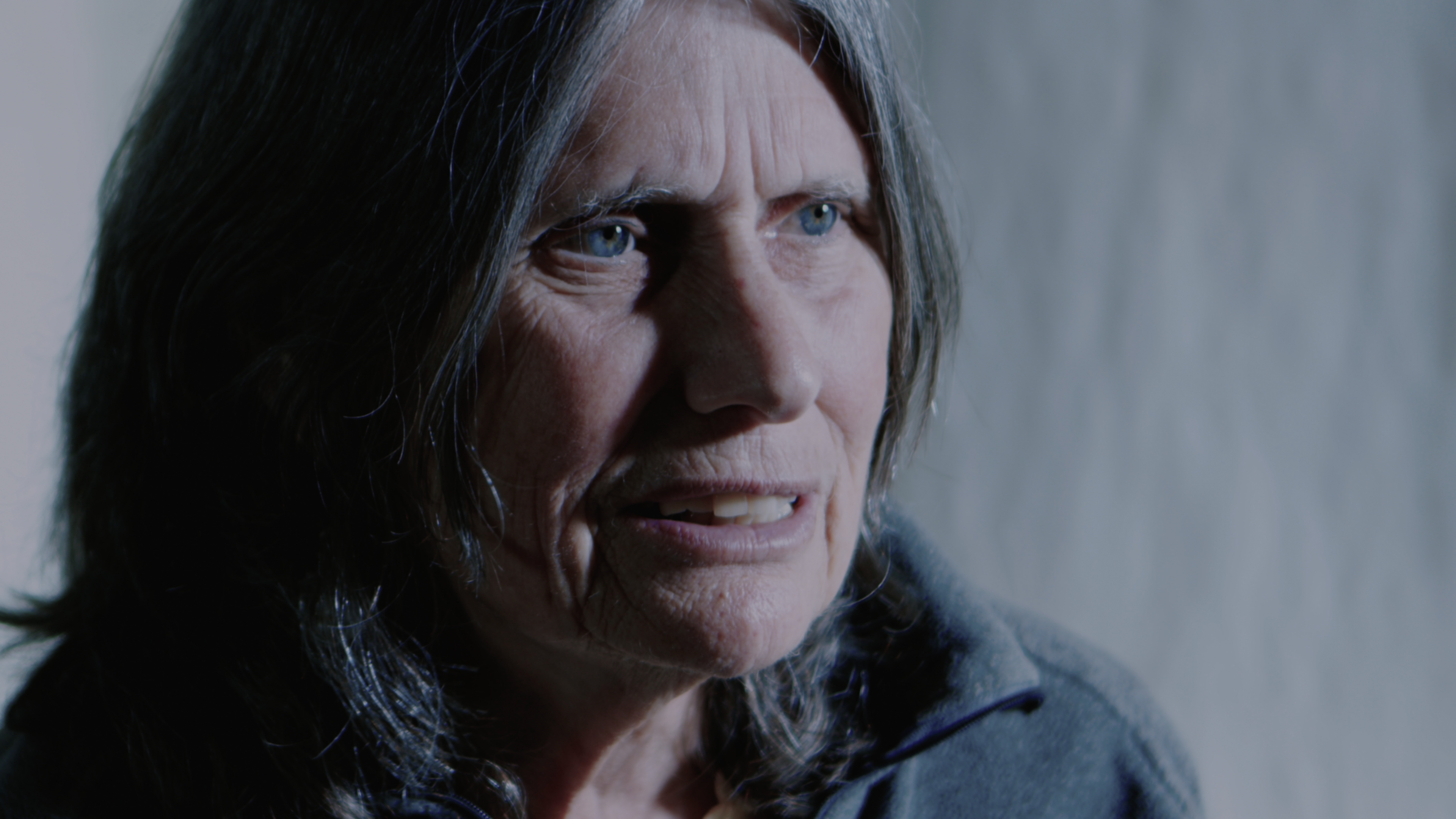 An older woman with long gray hair and blue eyes looks slightly to the side with a thoughtful expression against a plain background.