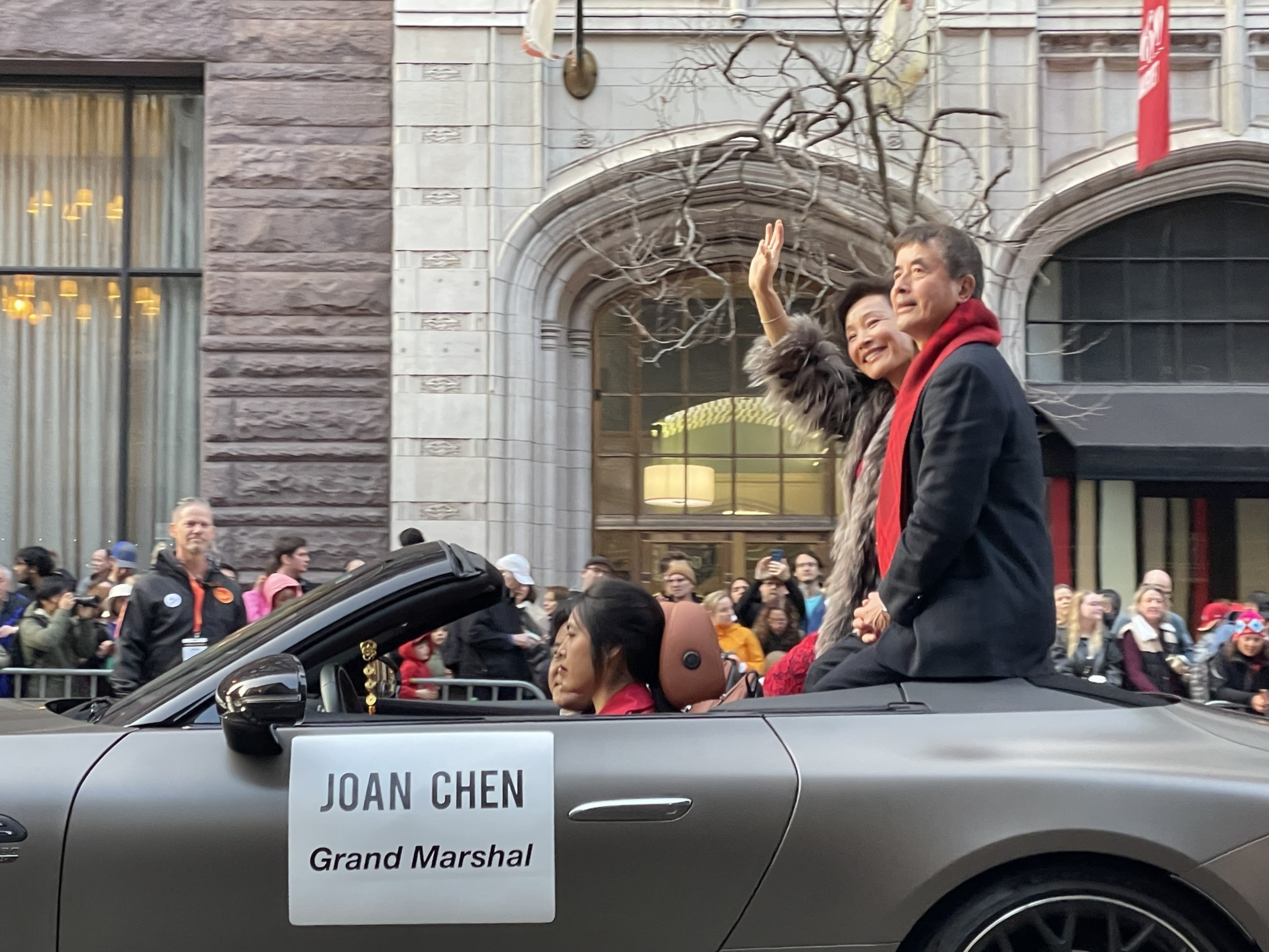 A smiling couple, with the woman waving, rides in a convertible labeled &quot;Joan Chen Grand Marshal&quot; during a parade, surrounded by spectators.
